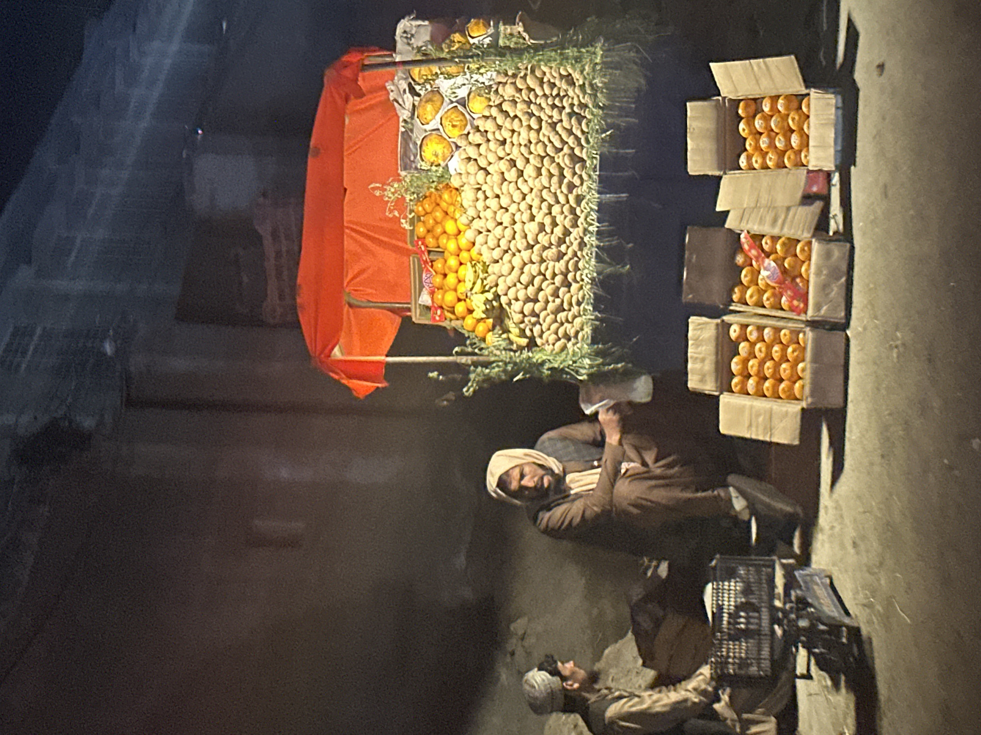 The scene depicts a dimly lit outdoor street corner in Quetta City Tehsil, Pakistan, at night. A fruit vendor's stall dominates the view, laden with oranges, potatoes, and other produce, arranged both on the display and in boxes nearby. Two men are visible, likely the vendors; one sits near the stall, while the other rests on the ground nearby. A scale and crate are situated near the men. The area is dark and appears to be a quiet commercial or residential street.