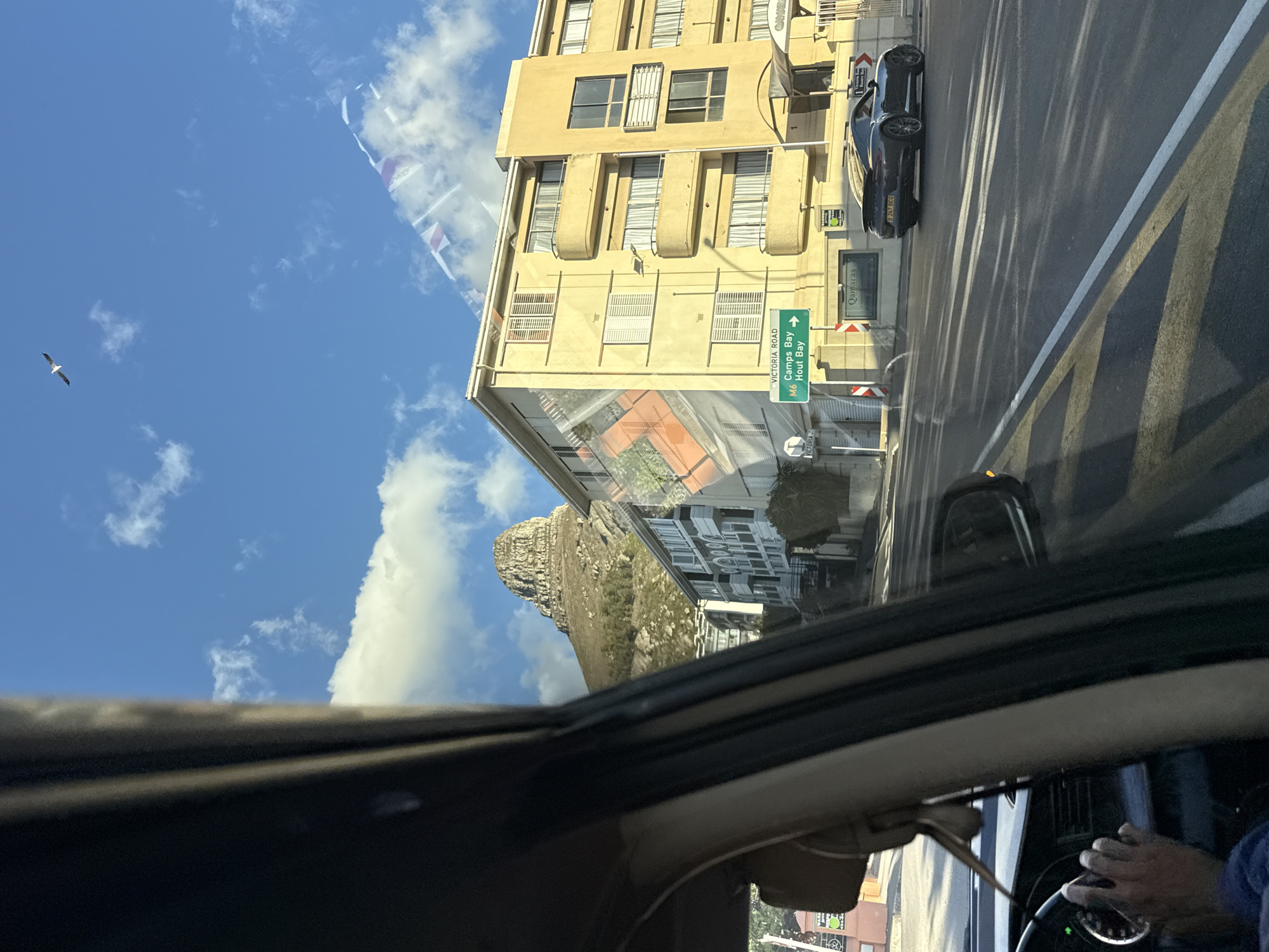 The image, taken from inside a vehicle, shows a view of Cape Town, South Africa. The driver's hands are visible on the steering wheel. Outside, there is a yellow building with multiple windows and a street sign indicating the direction to Camps Bay and Hout Bay. A dark-colored car is parked beside the building. In the background, a mountain is visible, partially obscured by clouds. The sky is blue with scattered white clouds. A bird is seen flying in the sky. Pennants strung across the street are also visible.