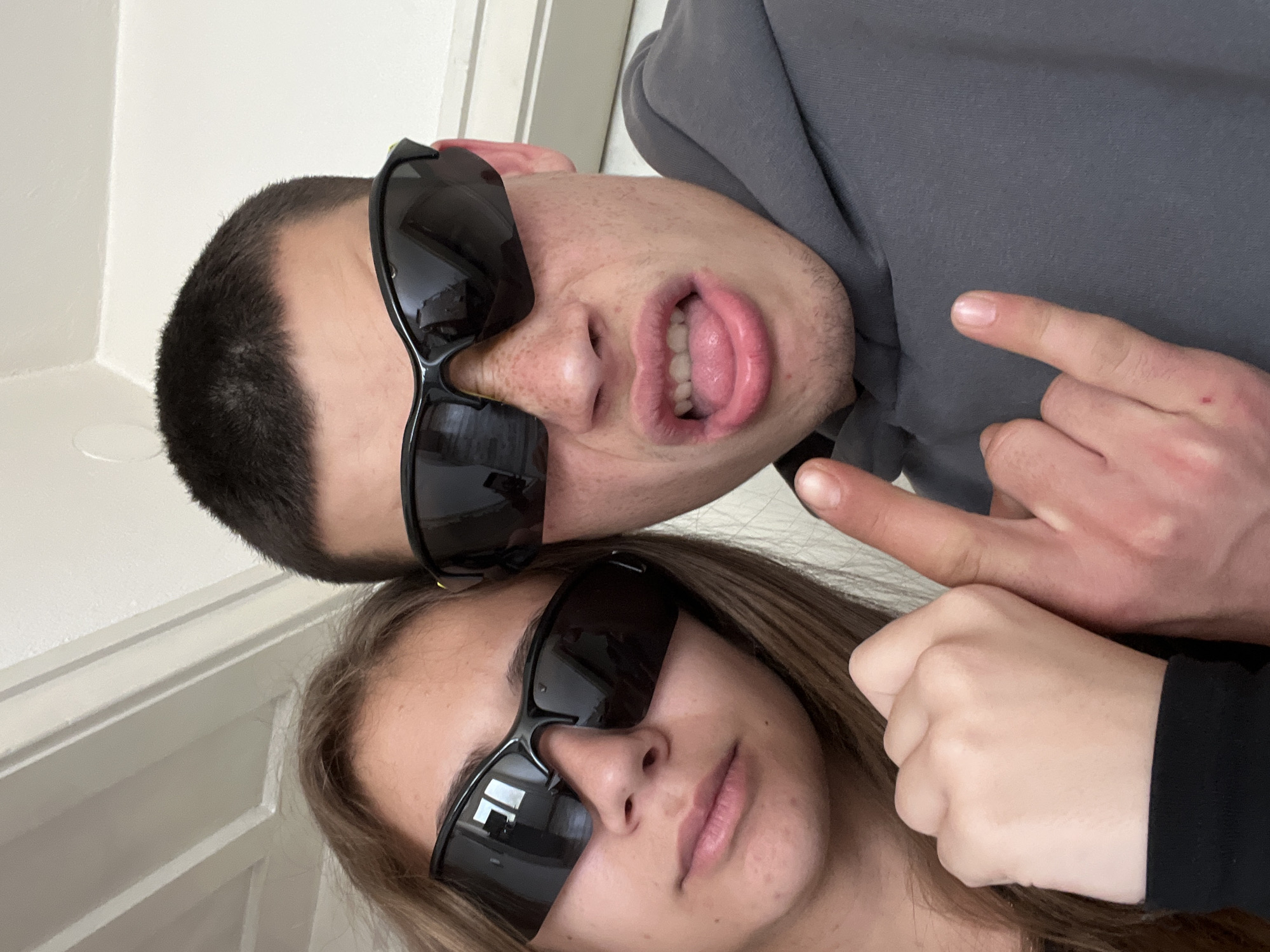 The image depicts two individuals wearing sunglasses, one male and one female, indoors. The male is positioned above the female, with a gray hooded sweatshirt visible. His mouth is open, exposing his teeth and tongue. The female's face is oriented below his. A hand, presumably belonging to one of the subjects, is making a hand gesture. The location is not identifiable as Karavelovo, Bulgaria, based on the visual information. The background includes a white wall and a door frame.