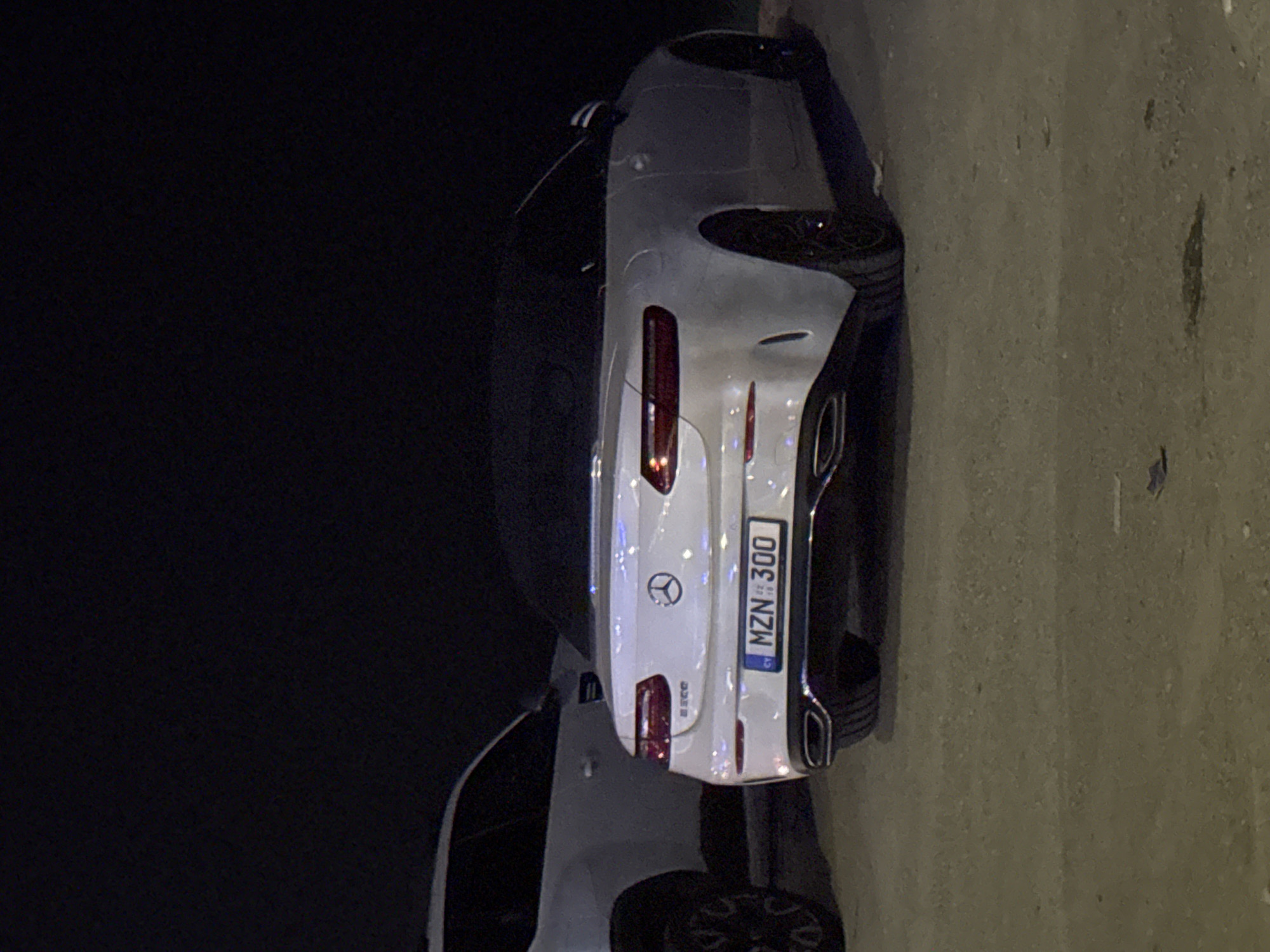The image shows the rear of a white Mercedes-Benz E-Class convertible parked at night. The license plate reads "MZN 300" and has a Cyprus country code. To the left, partially visible, is a white SUV. The ground appears to be a dirt or gravel surface, and the overall lighting suggests an outdoor urban or suburban environment, consistent with Germasogeia Municipality, Cyprus. No people or actions are depicted.