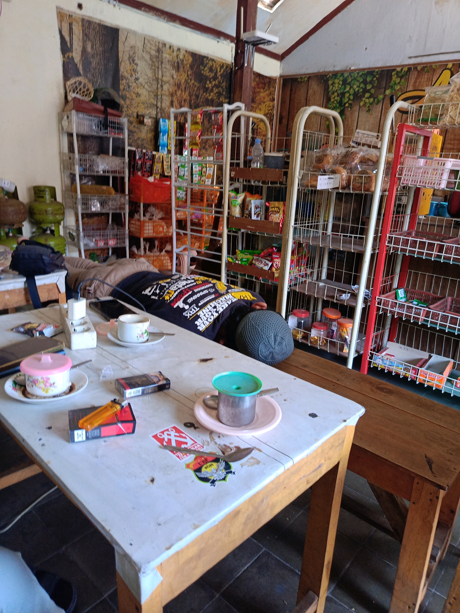 The image depicts a small shop interior in Kebonagung, Indonesia. A person is lying down on a table, appearing to be asleep. The table is cluttered with various items, including a pack of cigarettes, a lighter, a cup of coffee, and a smartphone. Shelves filled with snacks and household items line the walls. The overall impression is of a casual, unpretentious establishment.