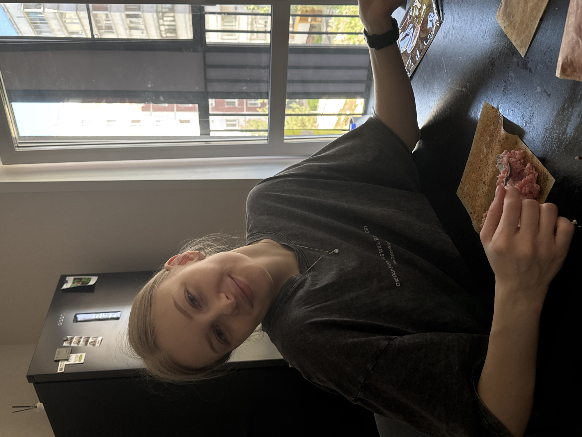 A young woman with blonde hair is in a kitchen, potentially in an apartment, preparing food. She is wearing a dark grey t-shirt and has a silver necklace. She is using a utensil to place a portion of raw meat onto a square piece of material, likely pastry or dough. On the table, there is another piece of the same material and a tablet device. Behind her is a black refrigerator with attached magnets. A window in the background reveals a cityscape with multiple buildings, suggesting an urban location, potentially in Perm, Russia, based on the location of the request. The blinds of the window are partly drawn. She is looking at the camera.