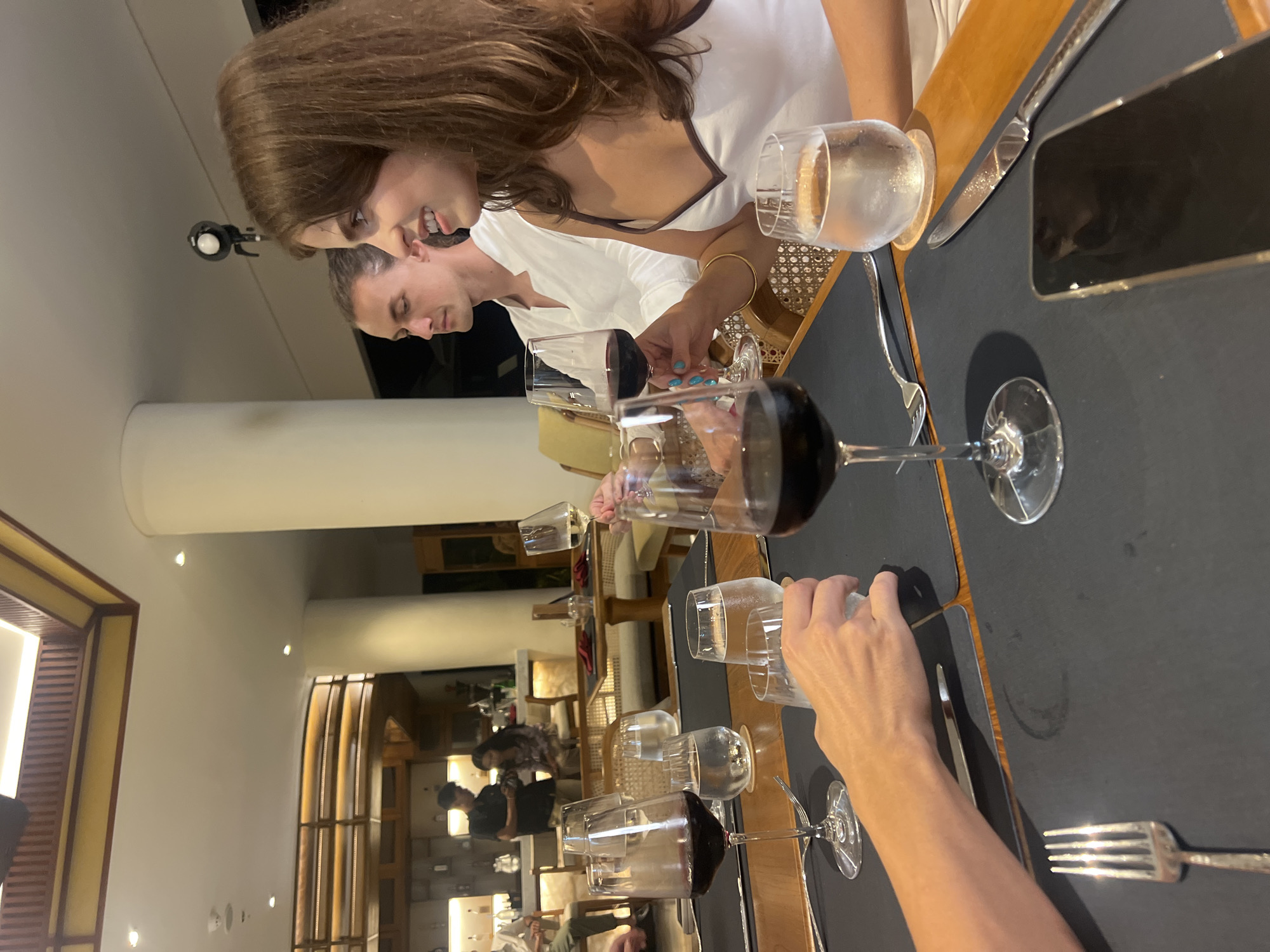 This media file shows a group of people seated at a table, likely in a restaurant, in Tegallalang, Indonesia. Two women are prominent in the foreground, with their faces partially visible. One woman, with long brown hair, is looking down and smiling. Next to her, another person, possibly a man, is also looking down. The table is set with multiple wine glasses, some containing red wine and others water, and cutlery. A phone is visible on the table, along with a knife and fork. In the background, other diners can be seen at different tables, and the setting appears to be an indoor dining area with some decorative wooden elements. The lighting suggests it is evening. The overall mood seems relaxed and social, as if people are enjoying a meal together.