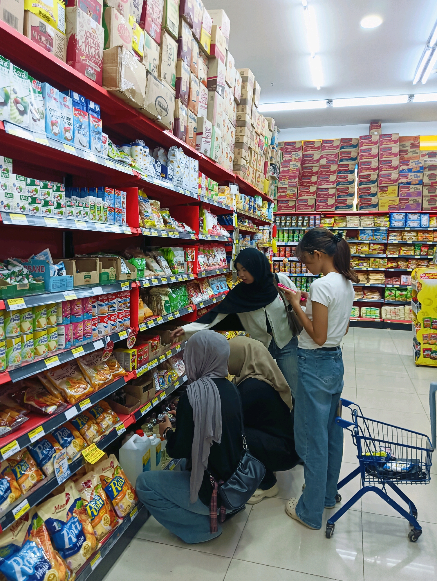 In a brightly lit supermarket aisle in Batam, Indonesia, three women are browsing the shelves. A woman wearing a black hijab and a light-colored cardigan is looking at her phone, while a woman in a white t-shirt and jeans stands next to her, also holding a phone. In the foreground, a woman in a grey hijab and black abaya is crouched down, examining items on a lower shelf. Adjacent to her is a small blue shopping cart. The shelves are fully stocked with various packaged goods, including what appear to be food items, beverages, and household supplies. The overall environment suggests a typical retail setting.