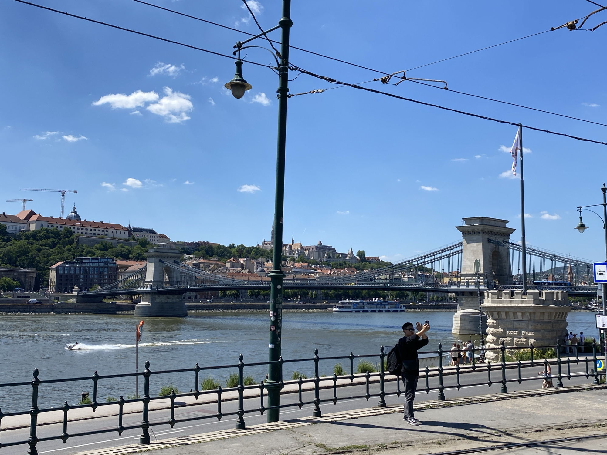 The image shows a daytime scene in Budapest, Hungary, with the Széchenyi Chain Bridge prominently featured. A pedestrian stands taking a photograph of the bridge with their smartphone. Behind them, other people are standing by the railing. The Danube River flows in the foreground, with a motorboat on the water. Across the river is Buda Castle Hill, with the castle visible at the top. The sky is blue with scattered clouds. Overhead are tram lines and lampposts. A flag on a pole is also visible on the right side of the image.
