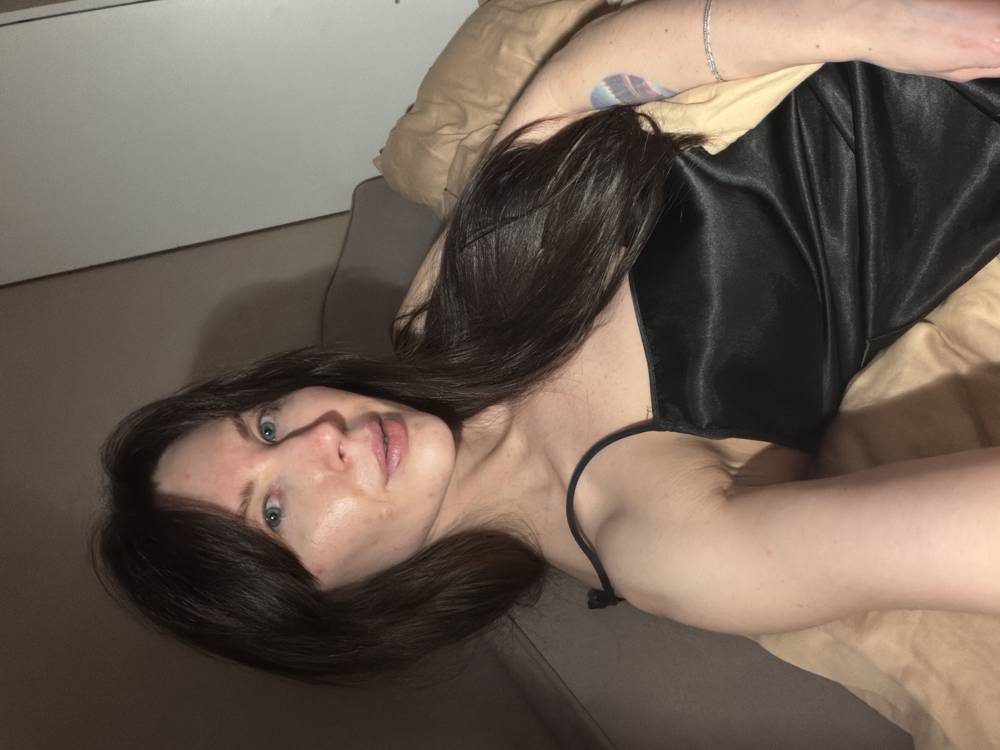 A woman with long dark hair and fair skin lies on a bed or sofa. She is wearing a black satin top with spaghetti straps. The background suggests an interior setting with beige bedding and a neutral-colored wall or furniture. The location and city of Perm, Russia cannot be determined from the provided image.