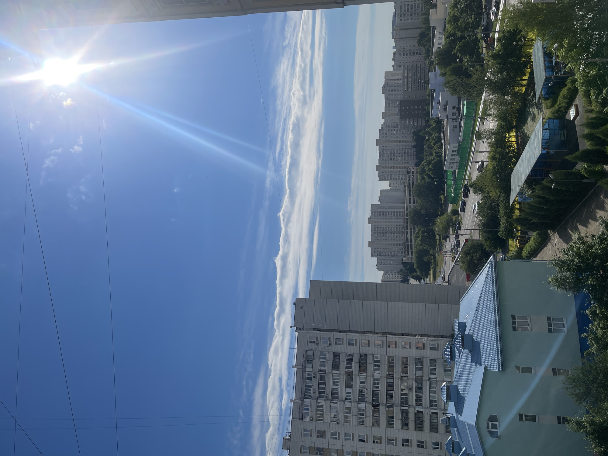 The image depicts a panoramic view of a residential area under a bright, sunny sky with scattered clouds and strong sun glare in Moscow, Russia. In the foreground, a light teal building with a blue tiled roof and multiple windows is visible. To the left, a tall, multi-story residential apartment building with numerous windows dominates the scene. Further in the background, a dense cluster of similar high-rise apartment buildings stretches across the horizon, indicative of a typical Moscow residential district. A road with several cars is visible between the buildings, bordered by trees and green fencing. The scene appears to be taken from a high vantage point, possibly another apartment building, looking out over the urban landscape.