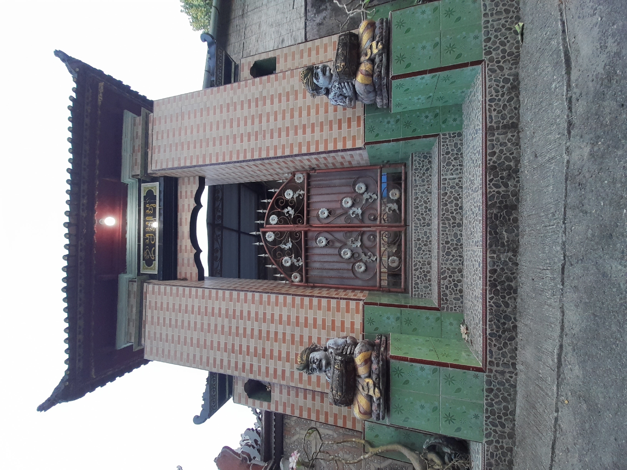 The image displays a brightly lit, ornate building entrance, likely a temple, shrine, or traditional structure, viewed from a slightly low angle. The central feature is a large, dark brown metal gate, intricately designed with various circular floral patterns and pointed decorative elements along the top.
Two substantial pillars, covered in light brown rectangular tiles, frame the gate. These pillars support a prominent roof structure with a multi-tiered, upward-curving edge, typical of East or Southeast Asian architectural styles. Above the gate, situated beneath the roof, is a black sign with a gold border featuring text in an unidentifiable Asian script. A single light fixture is visible and illuminated above this sign.
On either side of the entrance, positioned on raised platforms, are two statues. These guardian-like figures are seated or kneeling, depicted with detailed headwear and robes, and appear to be made of a grey stone or concrete material with noticeable gold accents. Steps and platforms leading up to the gate are covered with patterned tiles: the lower steps feature green square tiles with light floral designs, while the vertical risers and some platform sections are adorned with small, dark, pebble-like tiles. The ground to the right of the steps is a plain grey concrete surface. The clear and even lighting suggests it is daytime. No activity is depicted; the image focuses on the architectural details and decorative elements of the entrance.