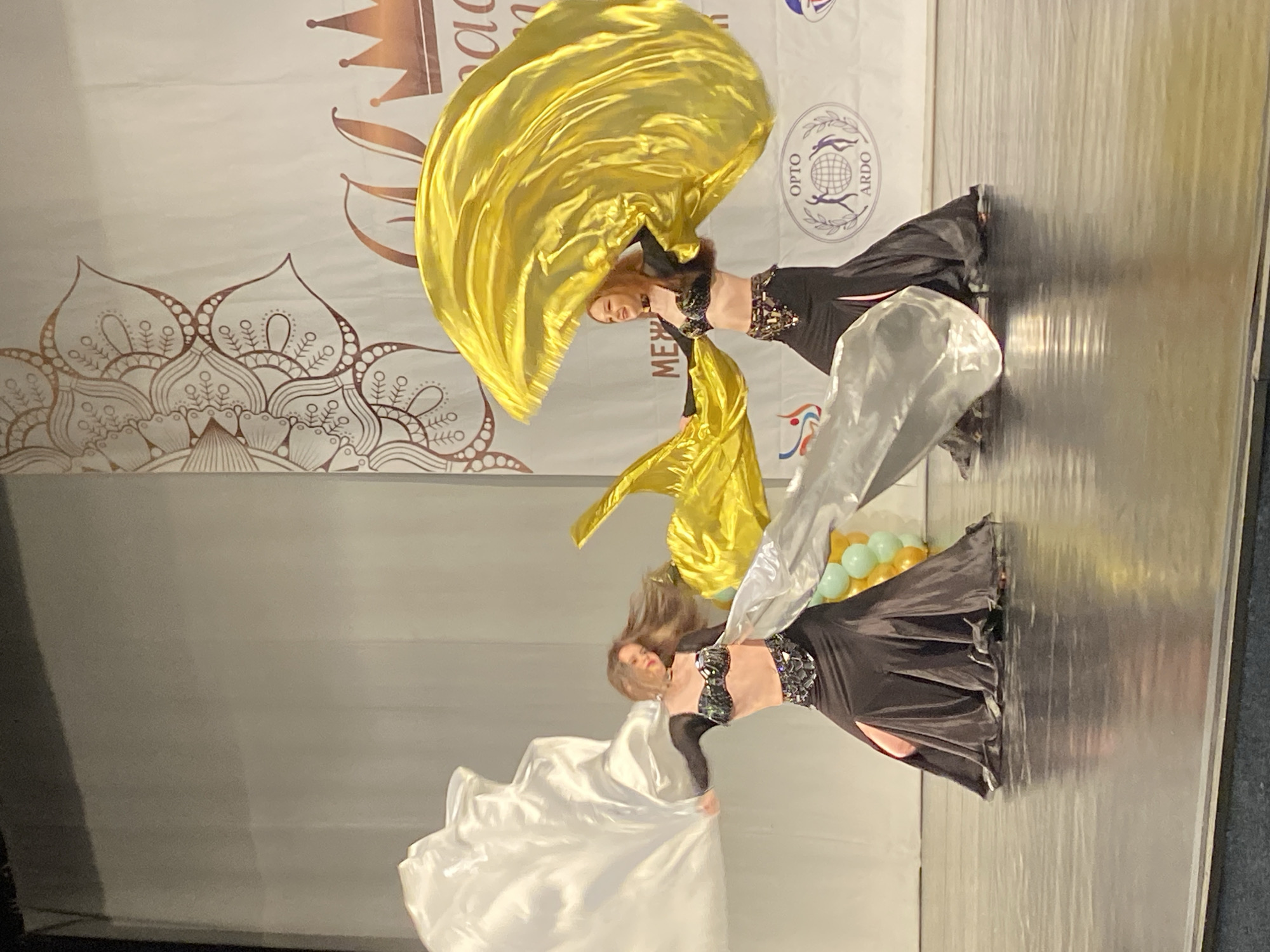 Two women are performing belly dancing on a stage. The woman on the left is holding a large white silk veil, while the woman on the right is holding a large gold silk veil. Both women are wearing black dance costumes. The background includes a large banner with a crown graphic and the words "MEZ..." visible. There is also a logo that includes the words "OPTO" and "ARDO" visible on the banner. There are balloons in the background. The dance appears to be taking place in Izhevsk, Russia.