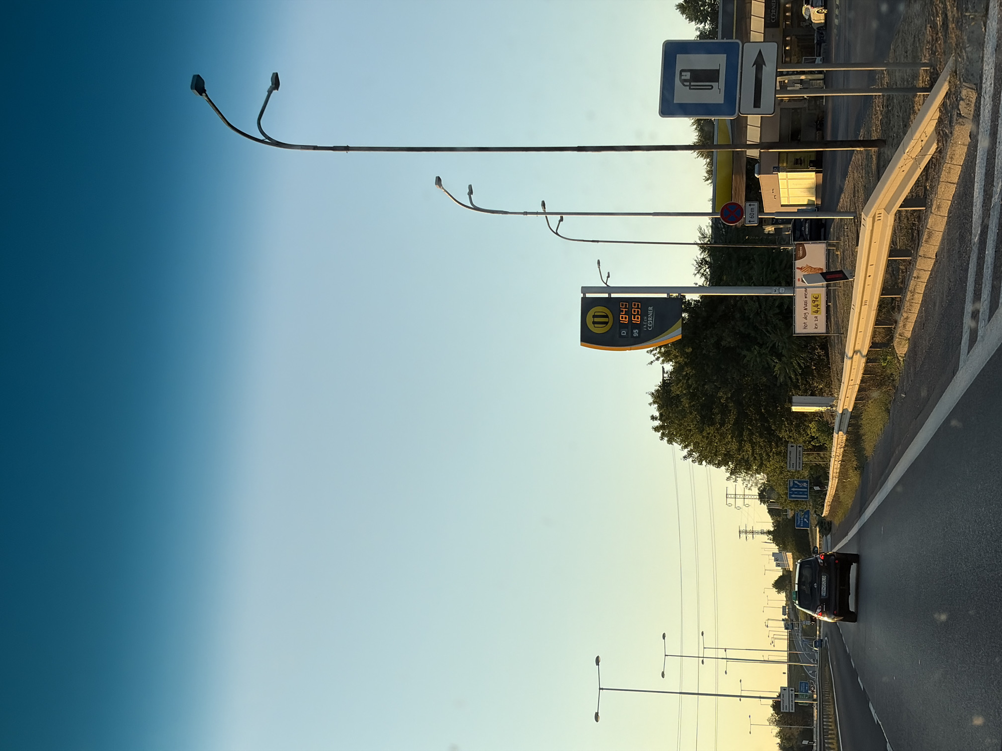 The image shows a highway scene with a car driving away from the camera, heading into the distance. On the right side of the road, there is a gas station with a prominent sign displaying fuel prices. The sky is clear with a gradient of blue to light yellow, suggesting either sunrise or sunset. Streetlights are visible along the highway. The location is identified as the city of Nitra, Slovakia, though specific landmarks confirming this are not readily apparent beyond the general highway infrastructure and gas station design.
