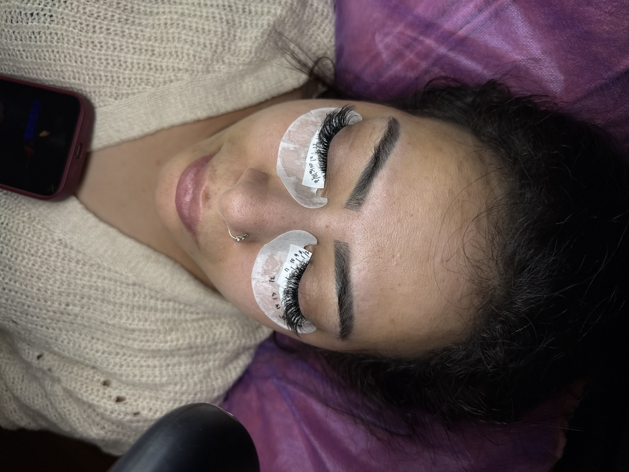 A person is lying down with their eyes closed, receiving eyelash extensions. The person is wearing a light-colored, knitted sweater and has a nose ring. Underneath the eyes, white pads with markings are placed to protect the skin and separate the lashes. The person's hair is dark and long, spread out around their head. The background appears to be a purple textured surface, possibly a blanket or treatment table. The scene suggests a beauty salon or spa environment. There is no information available about the location in Pamplona, Spain.