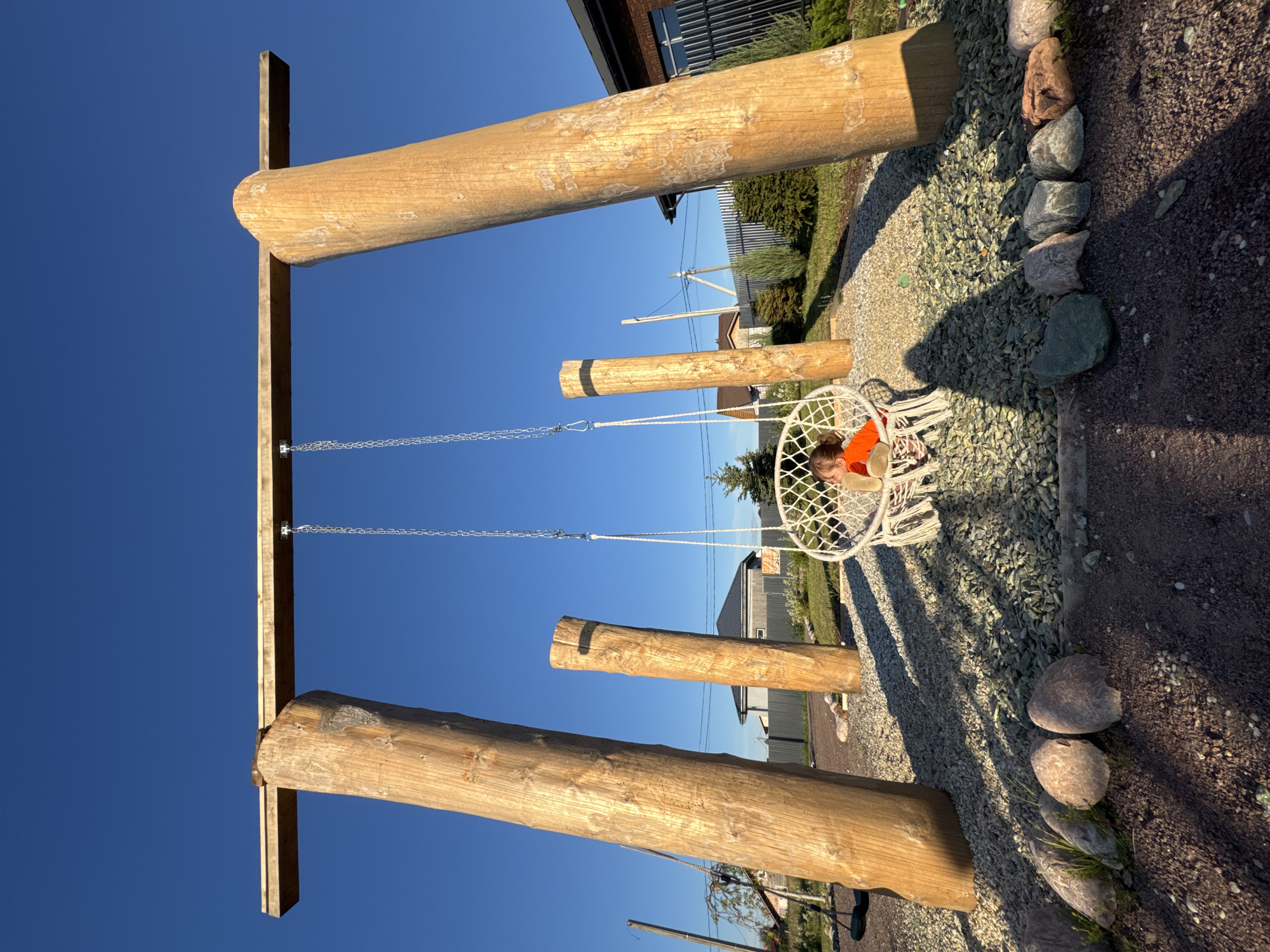 In Kempelevo, Russia, a child is seated in a white, macrame swing suspended from a large, wooden swing set. The swing set is constructed from four substantial, rough-hewn logs, two of which form the main uprights supporting a horizontal beam. The ground beneath the swing set is covered in a layer of small, green and grey gravel. To the left of the swing set, another log stands vertically, and further back, a smaller, dark-colored swing is visible. The background reveals a suburban environment with houses, fences, and trees under a clear blue sky. The lighting suggests it is late afternoon, with long shadows cast by the swing set and the child.