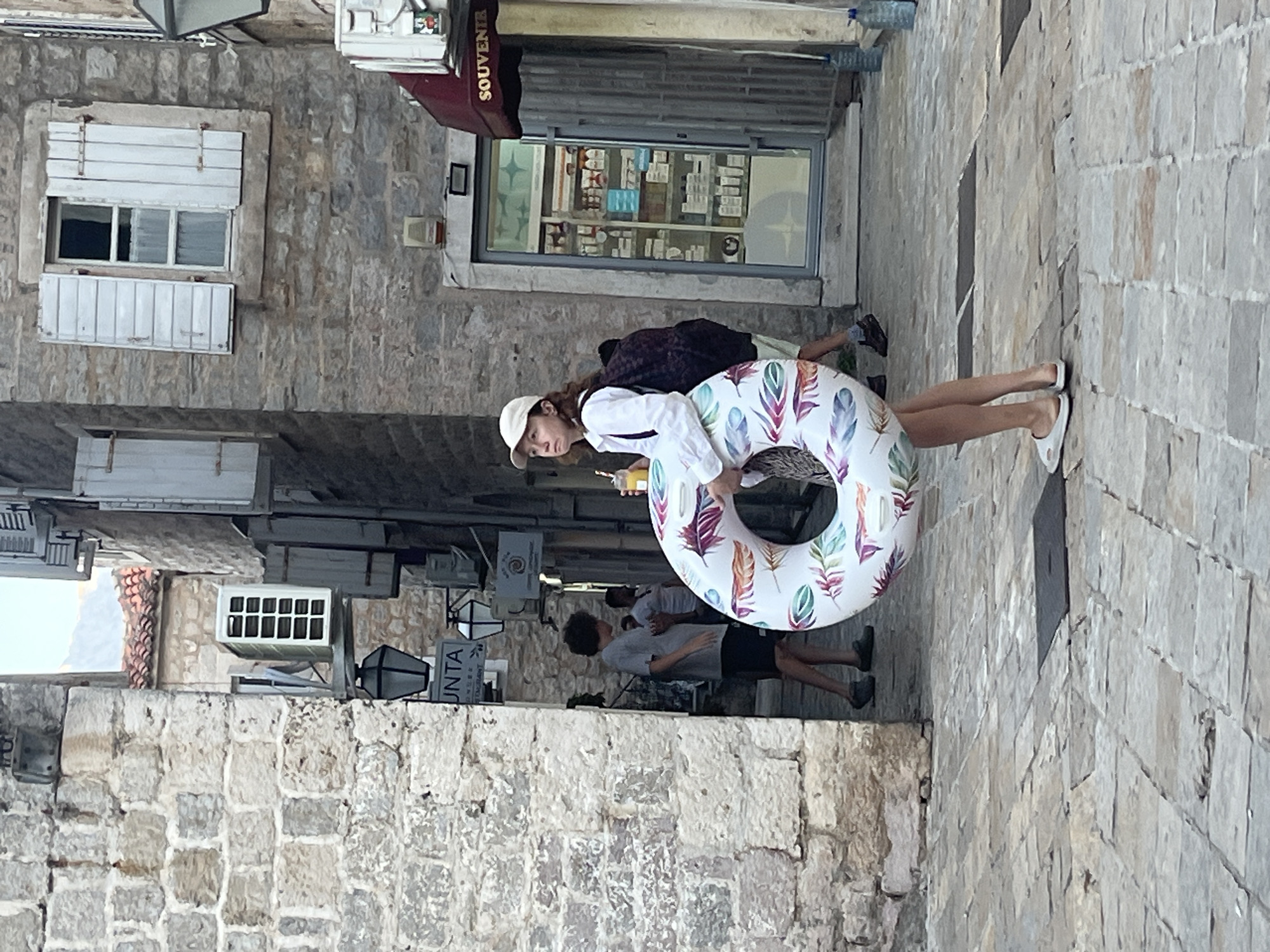A woman walks down a narrow, stone-paved street in Budva, Montenegro, carrying an inflatable ring decorated with feathers. She is wearing a white cap, a white shirt, and white slip-on shoes. A backpack is slung over her left shoulder, and she holds a drink in her right hand. Behind her, two young men walk in the same direction, one in a grey t-shirt and dark shorts, the other in a white t-shirt and dark shorts. The street is flanked by old stone buildings with visible air conditioning units and shutters. A souvenir shop with a display window is visible on the right side of the image. The ground is paved with rectangular stone slabs.