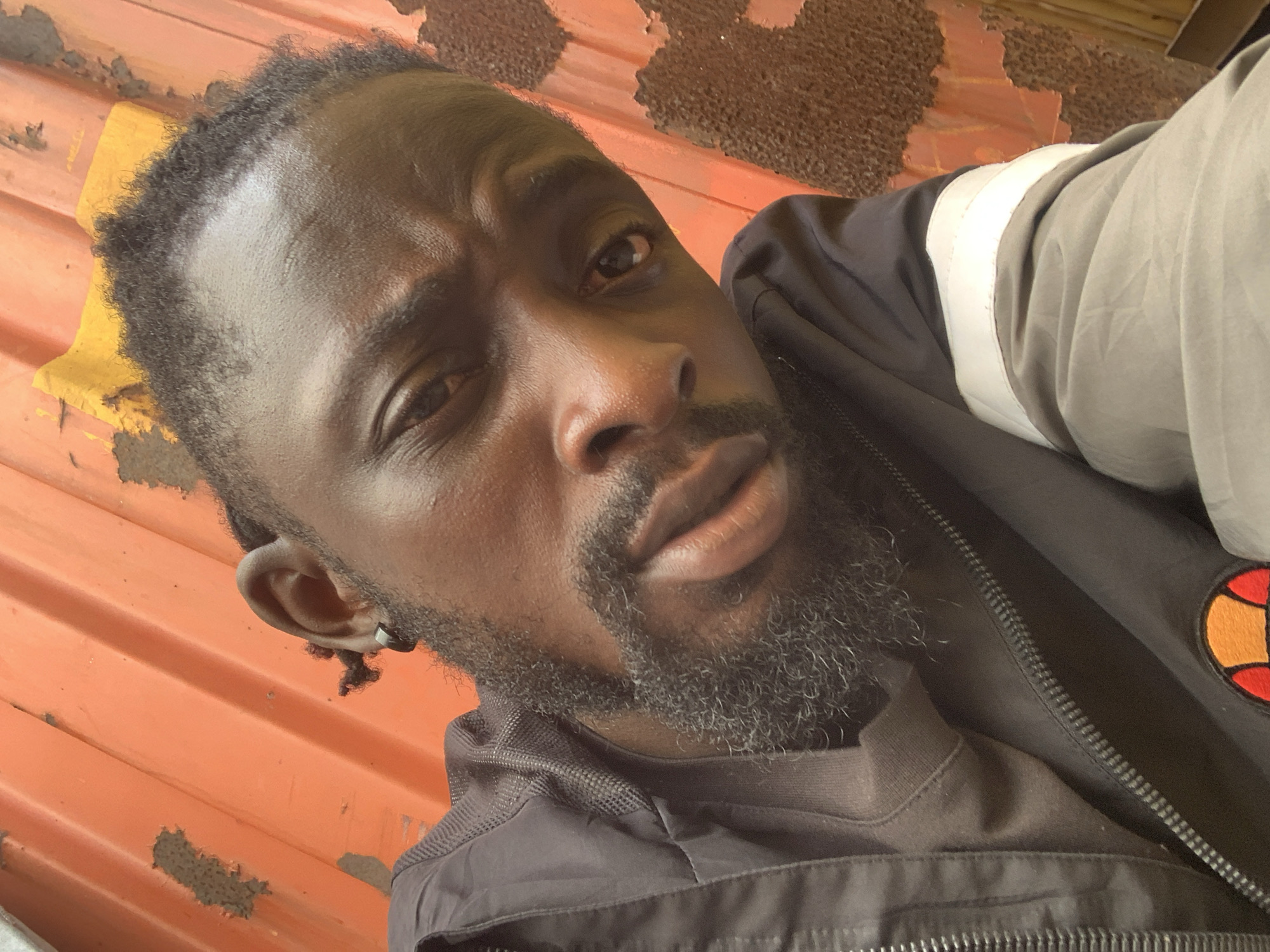 A medium-close-up portrait features a Black man with a serious and contemplative expression, looking directly at the camera. His head is slightly tilted. He has a dark skin tone, short textured hair along his hairline, and a full, dark beard and mustache showing some graying. A small silver hoop earring is visible in his left ear. His lips are full, and his brow is slightly furrowed, conveying an intense gaze.
He is wearing a dark, possibly black, zip-up jacket over a light grey shirt. The jacket has a distinctive white stripe across the upper arm and a colorful logo on the left chest/shoulder area, which appears to be the Ellesse brand emblem—a stylized 'e' or arch within a red, orange, and yellow circle.
The background consists of an orange-red corrugated metal surface, possibly a container wall or a building facade, showing significant signs of wear, rust, and peeling paint. This worn, industrial-style backdrop suggests an outdoor setting, potentially in an urban or commercial area consistent with Lekki, Nigeria. The lighting is bright and even, indicating daytime. No other individuals or discernible text are present in the image.