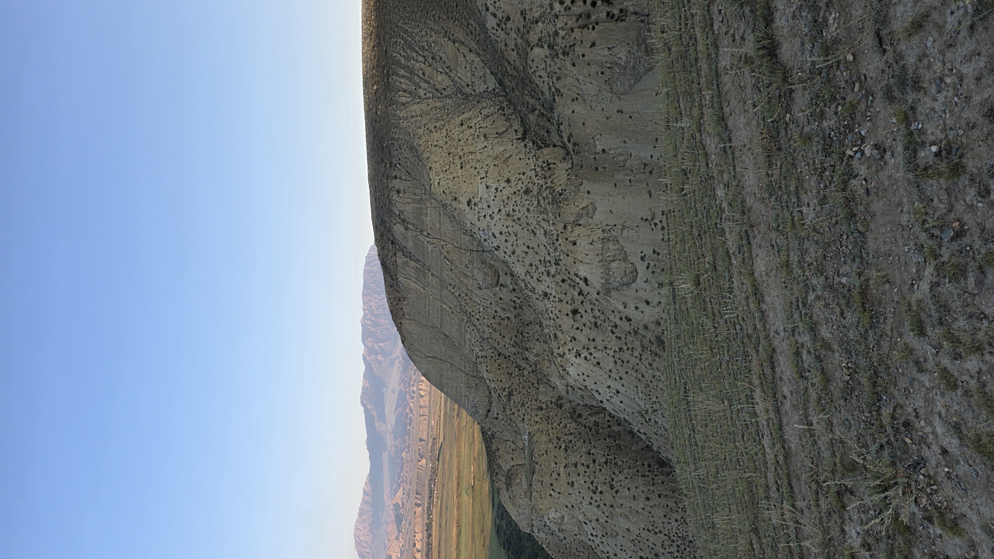The image depicts a vast, arid landscape with a clear, pale blue sky overhead. In the foreground, dry, sparse vegetation and soil cover a sloping terrain. A prominent, eroded hillside dominates the midground, characterized by its sandy, textured surface and scattered dark green shrubbery. The hillside exhibits visible layering and fissures, suggesting geological activity. In the distance, a valley stretches out, appearing greener and flatter, with what seems to be a small settlement or village nestled within it. Beyond the valley, a range of rugged, layered mountains extends towards the horizon, their peaks softened by atmospheric haze. The overall impression is one of a remote, natural environment with a sense of geological history and a subtle hint of human presence in the distant valley. The scene is consistent with the mountainous and valley terrain found in and around At-Bashi, Kyrgyzstan.