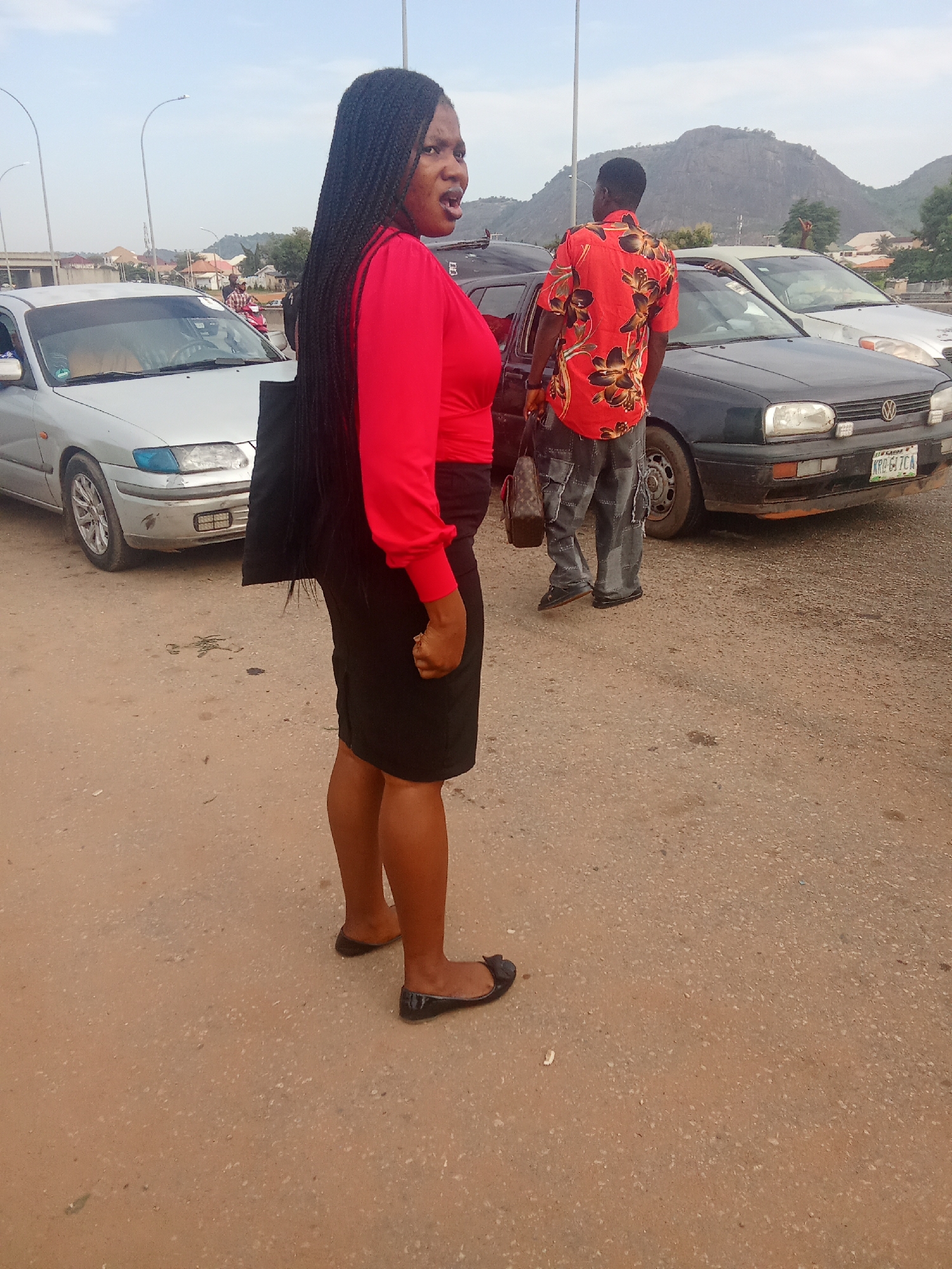 The image depicts an outdoor scene in Kagini, Nigeria, with a focus on three individuals and two vehicles. In the foreground, a Black woman with long braided hair, wearing a red long-sleeved top and a black skirt, stands facing left, with her mouth open as if speaking. She is carrying a black bag. Behind her and to the right, a Black man wearing a red and black floral-patterned shirt and patched denim pants stands facing away from the camera. He is looking towards a dark Volkswagen Golf car, and a portion of its front is visible. To the left of the woman, a silver sedan, possibly a Mazda, is parked. The background shows a hilly landscape with some buildings and scattered trees under a hazy sky. The ground is a mixture of dirt and gravel, with visible tire tracks and debris. Streetlights and utility poles are also present, suggesting an urban or semi-urban environment. The overall atmosphere appears to be during daylight hours.