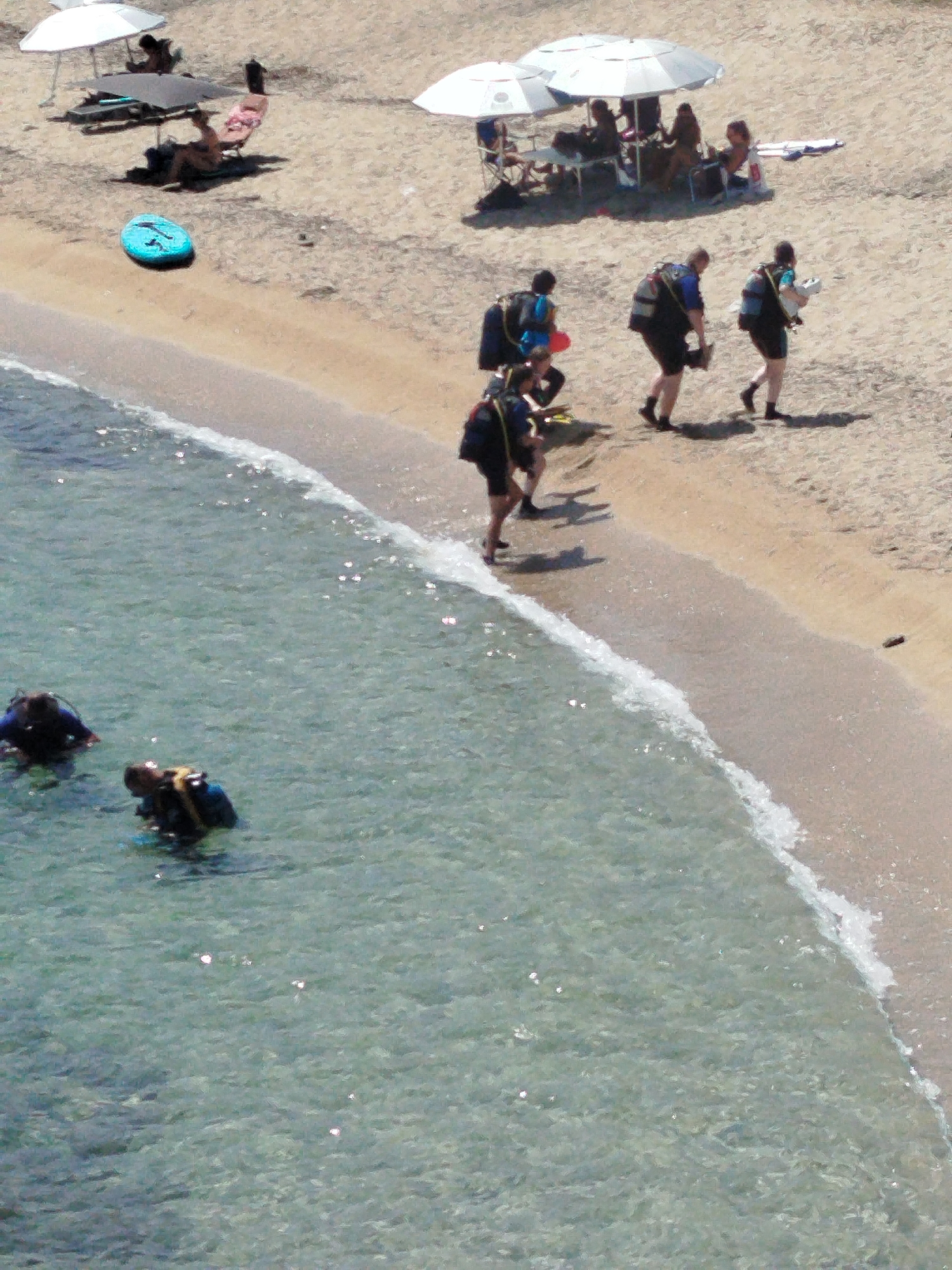 This is an aerial, high-angle, medium shot of a beach and the adjacent sea in Sithonia Municipal Unit, Greece. The sand is light brown, and the sea is clear and turquoise with small waves.
On the beach, several people are visible. In the foreground, two individuals wearing scuba diving gear are partially submerged in the water near the shore. They appear to be preparing for or concluding a dive. Further up the beach, a group of four people are walking away from the camera, also wearing scuba gear and carrying equipment. One person in this group is carrying a white object. Behind them, closer to the water's edge, a blue object resembling a bodyboard or surfboard rests on the sand. In the upper portion of the image, several people are relaxing on the beach under umbrellas. Some are seated in beach chairs, while others appear to be gathered around a table.
The environment is a sunny coastal scene with typical beach amenities like umbrellas and chairs. The water appears calm and suitable for water activities. The presence of scuba divers suggests this may be a popular spot for diving or snorkeling.