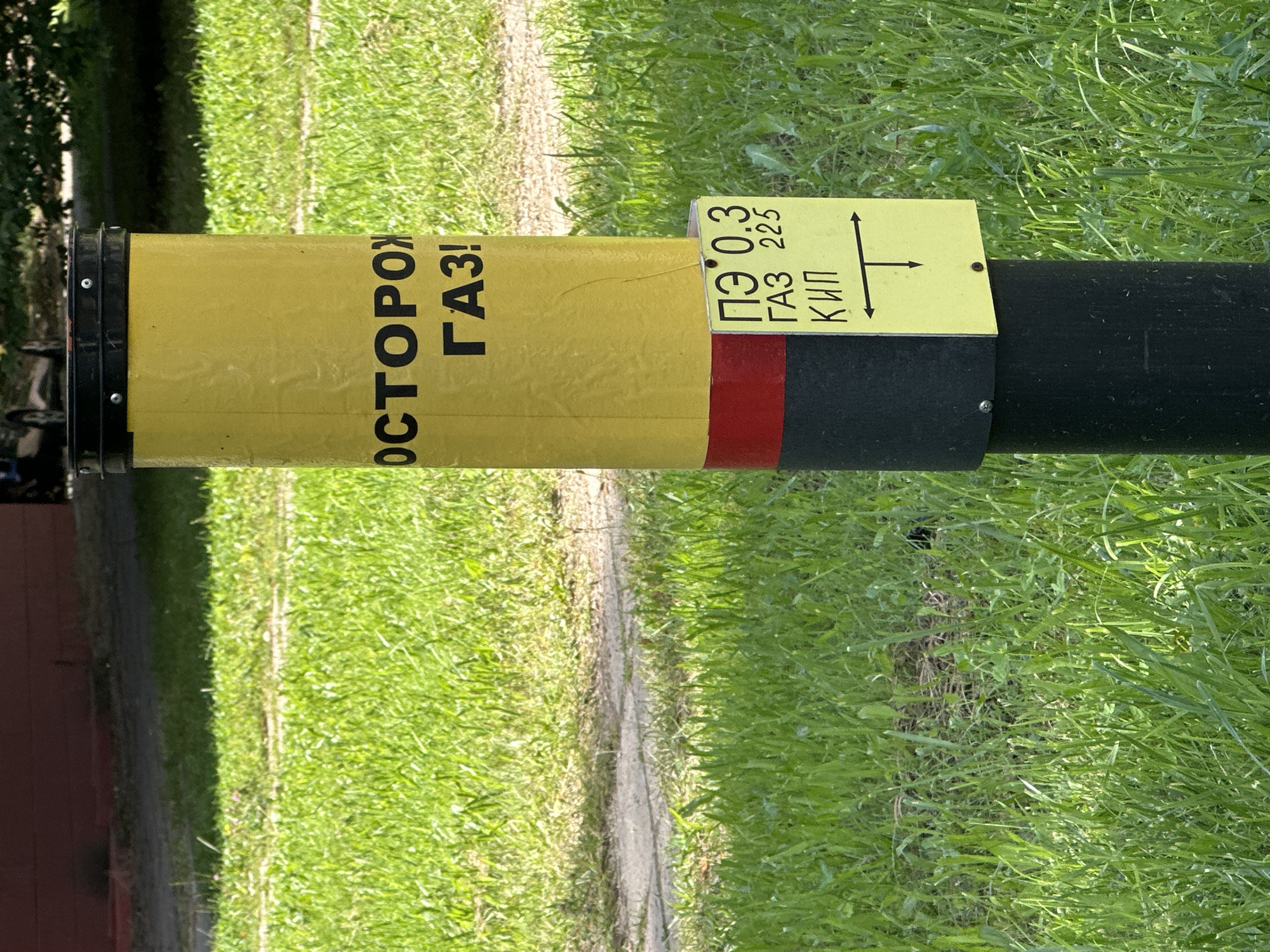 The image shows a yellow and black pole with red banding, situated in a grassy area. The pole has Russian text on it that reads "ОСТОРОЖНО ГАЗ!", which translates to "CAUTION GAS!". A smaller yellow sign is attached to the pole with more Russian text and symbols. The sign appears to indicate measurements or designations related to gas. The background consists of green grass and a blurry indistinct structure in the distance, possibly a building or fence. The scene appears to be outdoors, likely in a park or residential area, given the presence of grass and a walkway. No people are visible in the image. The image does not provide any specific information or context to determine its location in the city of Minsk, Belarus.