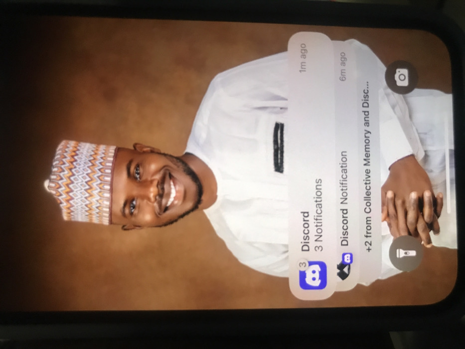 A portrait of a smiling man in a white outfit and patterned cap serves as a phone's wallpaper. Discord notifications are visible at the top. The first notification, marked as "1m ago," indicates three Discord notifications. The second notification, marked as "6m ago," is another Discord notification, mentioning "+2 from Collective Memory and Disc..." The phone's camera and flashlight icons are also visible on the right side of the screen.