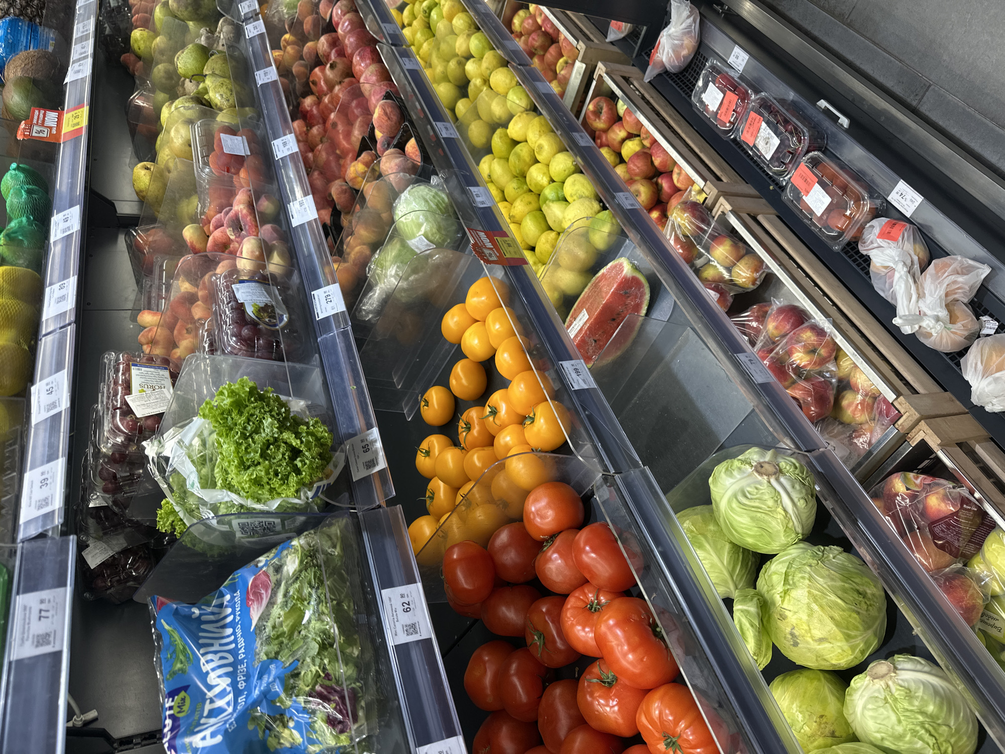 This is a photograph of a produce section in a supermarket in Ternopil, Ukraine. The shelves are stocked with a variety of fresh fruits and vegetables, including tomatoes, cabbage, lettuce, lemons, pears, peaches, cherries, and watermelons. The produce is arranged in plastic containers and bags, with price tags visible on some items. The lighting is bright and even, suggesting an indoor environment. There are no people visible in the image. The overall impression is one of abundance and freshness.