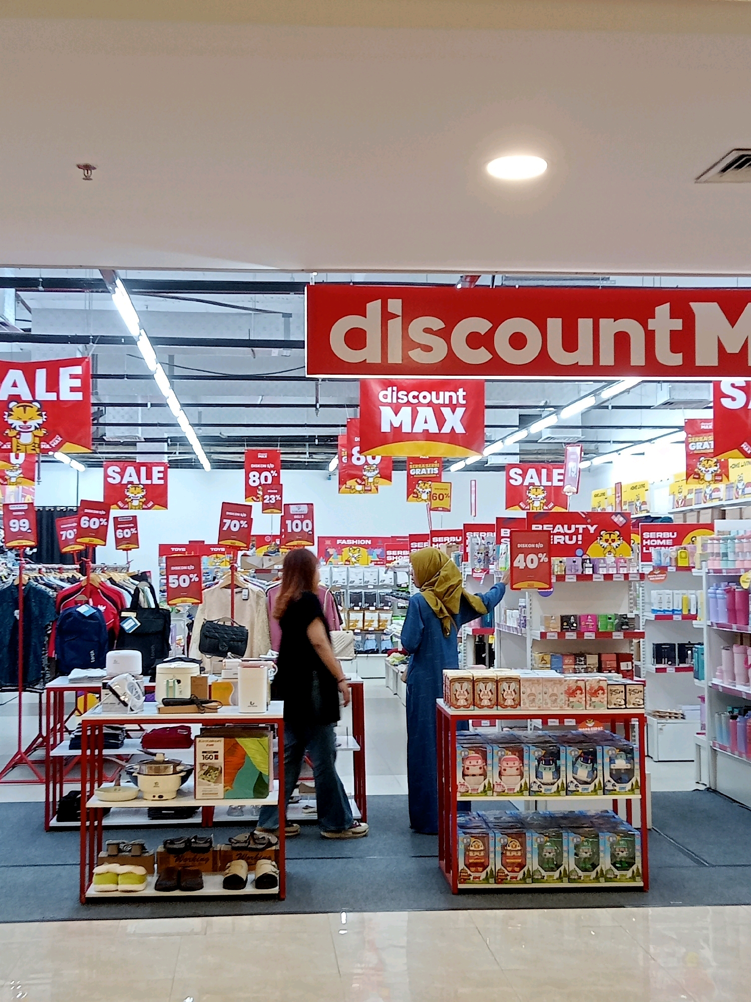 The image depicts an indoor retail environment, likely a department store or supermarket, with a focus on promotional signage and product displays. Several large red banners are prominently displayed overhead, featuring text such as "discount M" and "discount MAX," indicating ongoing sales or special offers. Smaller red signs with white text advertise percentages off, like "70%" and "60%", along with playful tiger graphics.
In the mid-ground, two individuals are visible. One person, wearing a black top and blue jeans, stands facing away from the camera, examining clothing racks. Another person, wearing a blue abaya and a yellow headscarf, is actively interacting with a display of beauty products, reaching towards a shelf. The background shows aisles of merchandise, including clothing on racks, shelves stocked with various products, and a prominent display of boxed items, possibly toys or small appliances, arranged on a red and white shelving unit. The overall lighting suggests a well-lit commercial space. The presence of merchandise and promotional materials points to a shopping or retail scenario. The style of the signage, specifically the use of "SERBU GRATIS" which means "Free Spree" or "Free Attack" in Indonesian, along with the tiger graphic which is often associated with Chinese New Year celebrations, suggests this store is located in Indonesia, and given the context of the prompt and the image content, it is likely in Cirebon.