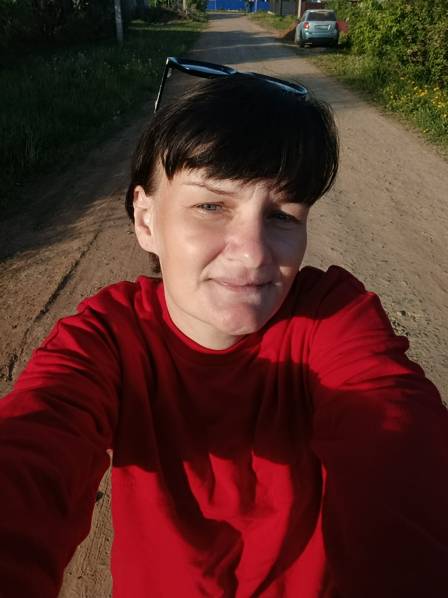 The image shows a person in a red shirt on a dirt road in Фоки, Russia. The person is taking a selfie, with sunglasses atop their head. The background shows greenery on either side of the road and a car parked further down the road. There is a building in the distance to the left.