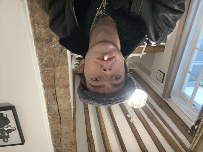 The image displays a close-up indoor portrait of a young male subject in a residential setting in Paris, France. The subject, positioned centrally, is a male with light skin and dark hair visible under a grey flat cap. He wears a black leather jacket with a black shearling collar and multiple silver-colored necklaces, including one with a cross pendant, over a dark shirt. A lit cigarette is held between his lips.
The environment exhibits architectural features characteristic of older Parisian buildings. The ceiling features exposed dark wooden beams, and a vertical wooden support beam is visible on the right side of the frame. The walls are painted white. On the left, a white-framed window allows natural light into the space, offering a view of an exterior building. A square, black-framed analog clock hangs on the wall near the window. A decorative light fixture with a woven shade is suspended from the ceiling. On the white wall to the right, a framed black and white photograph or print, possibly depicting musicians like The Beatles, is mounted. These details, particularly the exposed beams and window style, indicate an interior located within a historical building, commonly found in central districts of Paris.