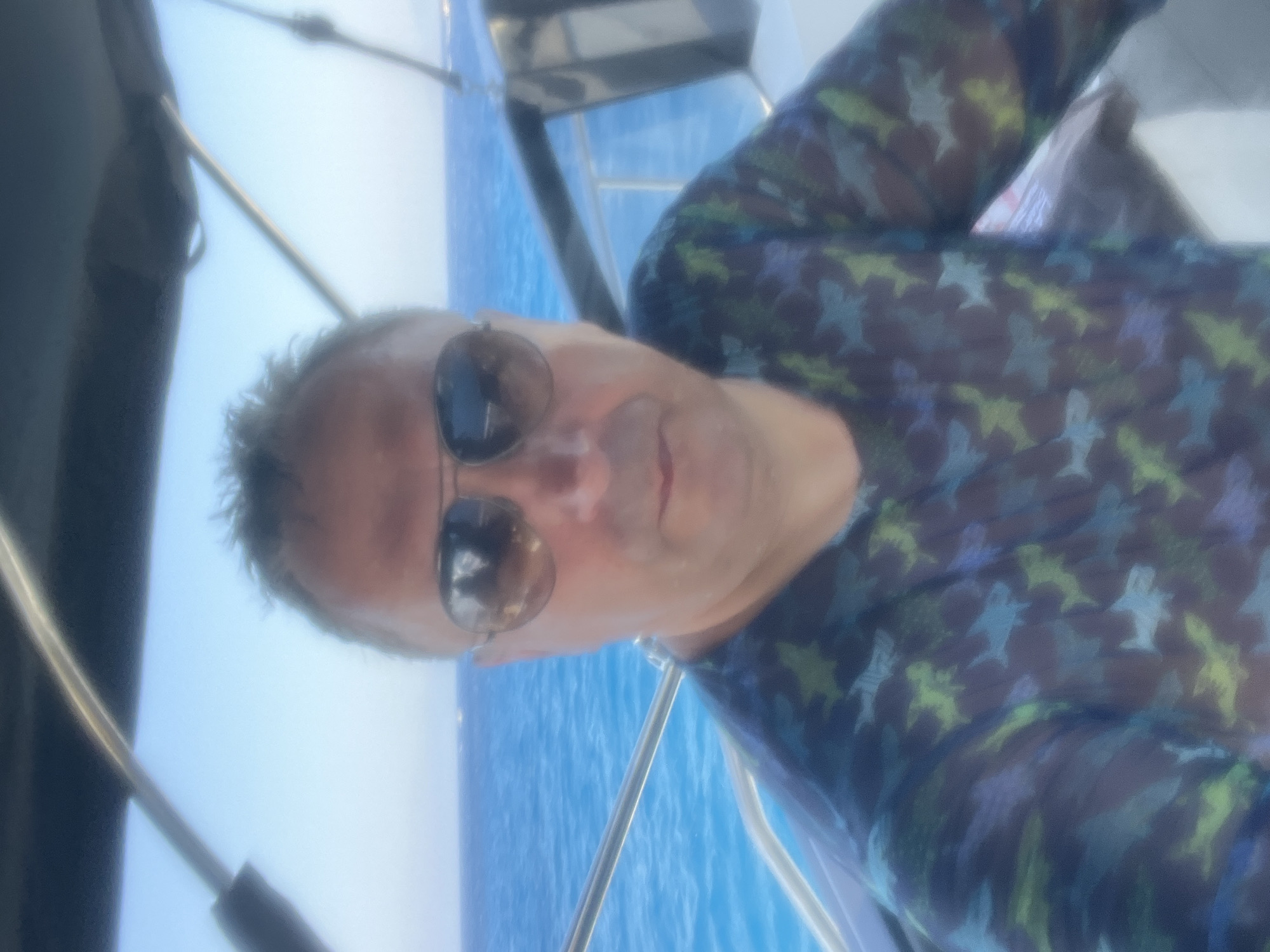 A man with short, graying hair, wearing aviator sunglasses and a dark blue long-sleeved shirt with a repeating pattern of light blue and green sharks, is pictured on a boat. He is looking directly at the camera. The background consists of a bright blue sky and clear, turquoise water. The boat's dark canopy is visible above the man's head, and a metal railing runs horizontally across the lower left of the frame. The image appears to have been taken on a sunny day. The location is the sea near Neo Chorio, Cyprus.