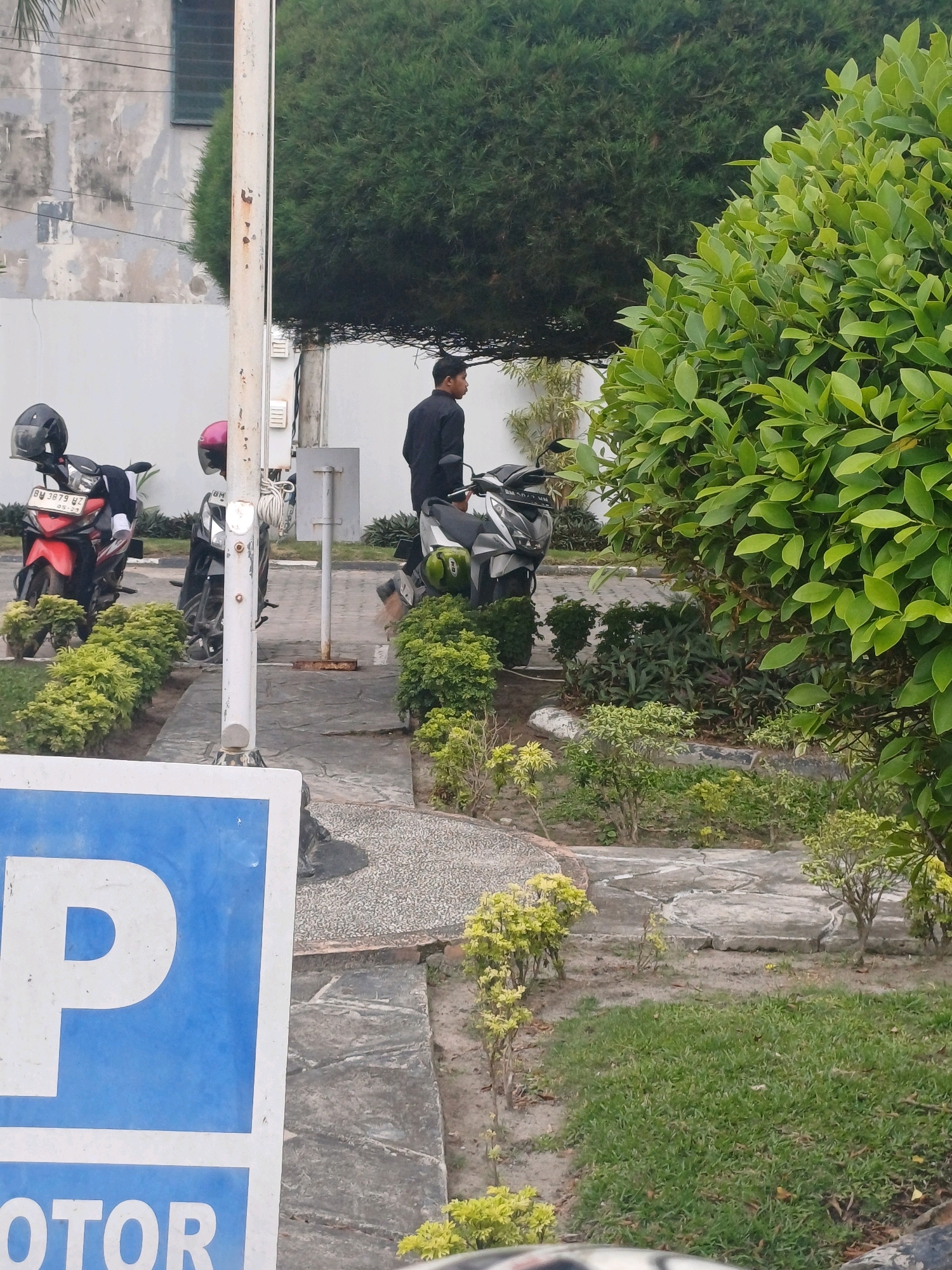 The image depicts a parking area with several motorcycles parked. A man in a black shirt is standing next to one of the motorcycles. A blue parking sign with a white "P" is visible in the foreground. The area is landscaped with various plants and trees, and a building with a textured wall is in the background. The scene appears to be outdoors in a city setting, possibly Dumai City, Indonesia, given the context.