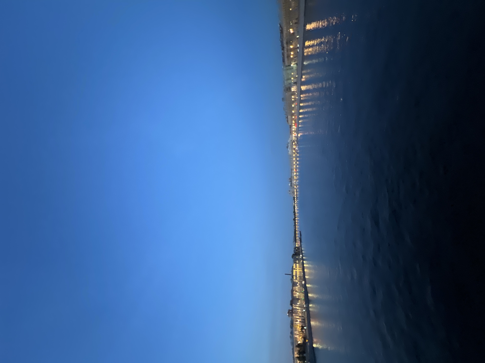 A wide, dark body of water, likely a river, dominates the lower half of the image. The water's surface is slightly rippled, reflecting the lights from buildings on the distant shore. The buildings form a long, continuous line along the embankment, illuminated by numerous warm-toned lights that create streaks of reflection on the water. The architecture appears to be historic, with a sense of grandeur. The sky above is a deep, clear blue, indicating either dawn or dusk. The overall atmosphere is serene and tranquil.