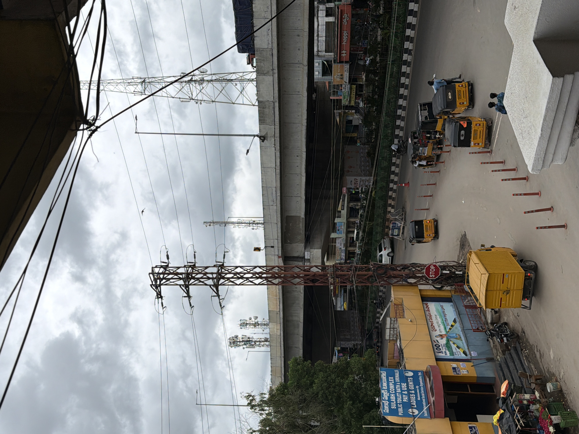 The image captures a cloudy day in Shamshabad, India, likely near a major intersection. In the foreground, a yellow auto-rickshaw truck is parked. To its left, a motorcycle is parked near the entrance of a yellow building labeled "SULABH COMPLEX PUBLIC TOILET BATH & URINALS PAY & USE LADIES & GENTS". A mural is painted on the side of this building. Further into the scene, an elevated highway crosses overhead, supported by concrete pillars. Several tall communication towers are visible on either side of the highway. On the road below the highway, a line of yellow auto-rickshaws and a few cars are present. Two men are interacting near one of the auto-rickshaws. The sky is overcast with heavy clouds, and electrical wires crisscross the upper portion of the frame.