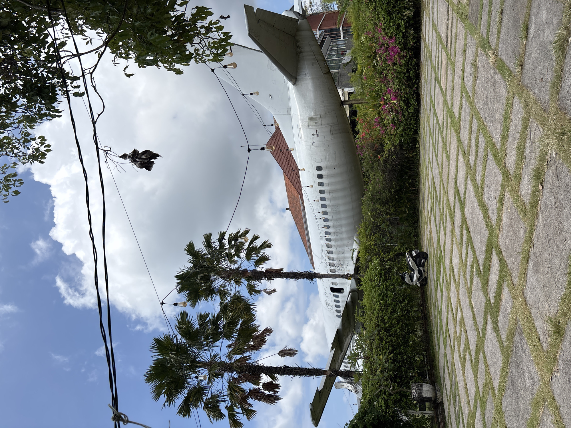The image shows a grounded airplane fuselage in Kuta, Indonesia, repurposed as part of a building or attraction. The tail section of the white plane is visible, along with several rows of windows. The structure has a red-tiled roof incorporated into its design. A paved walkway with grass growing in the gaps runs alongside the fuselage. Lush greenery and flowering bushes surround the structure. Palm trees are also present, with string lights suspended between them. Above, the sky is partly cloudy. Electrical wires run across the upper left quadrant of the frame, obscured by foliage. A parked motorbike is visible next to the walkway.