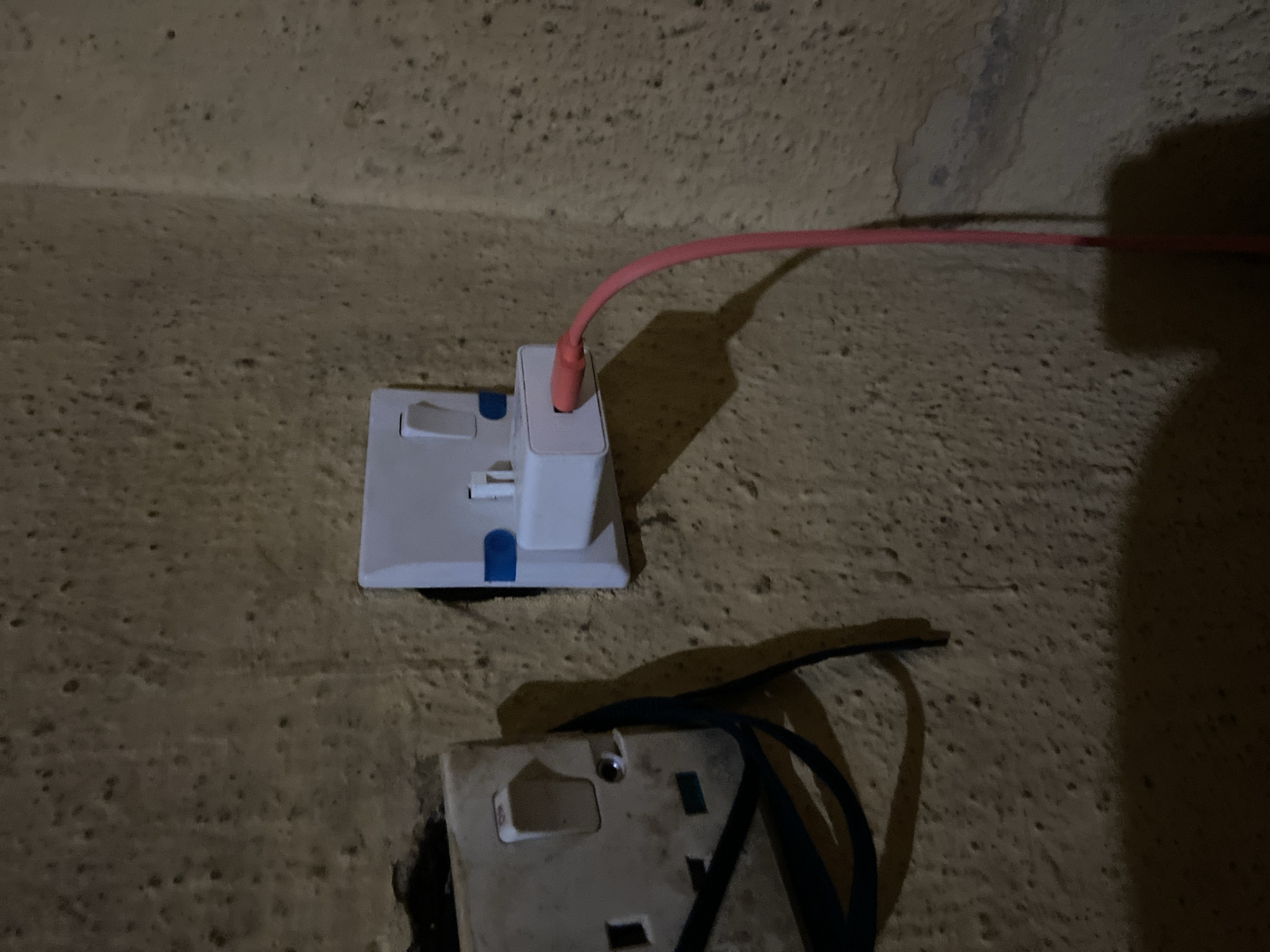 The image shows a close-up view of two electrical outlets on a textured, yellowish wall. On the left is a white three-pin socket with a toggle switch and some dark cables plugged in or attached. To the right of this is a white USB charger plugged into a wall socket. A pink charging cable extends from the USB charger downwards. The wall appears weathered and has a rough, uneven surface. Shadows are cast by the electrical fittings and the cables. The scene is indoors, likely in a residential or commercial building, given the presence of electrical outlets. The specific location in Jalingo, Nigeria, is not indicated by any visible landmarks or identifying features.