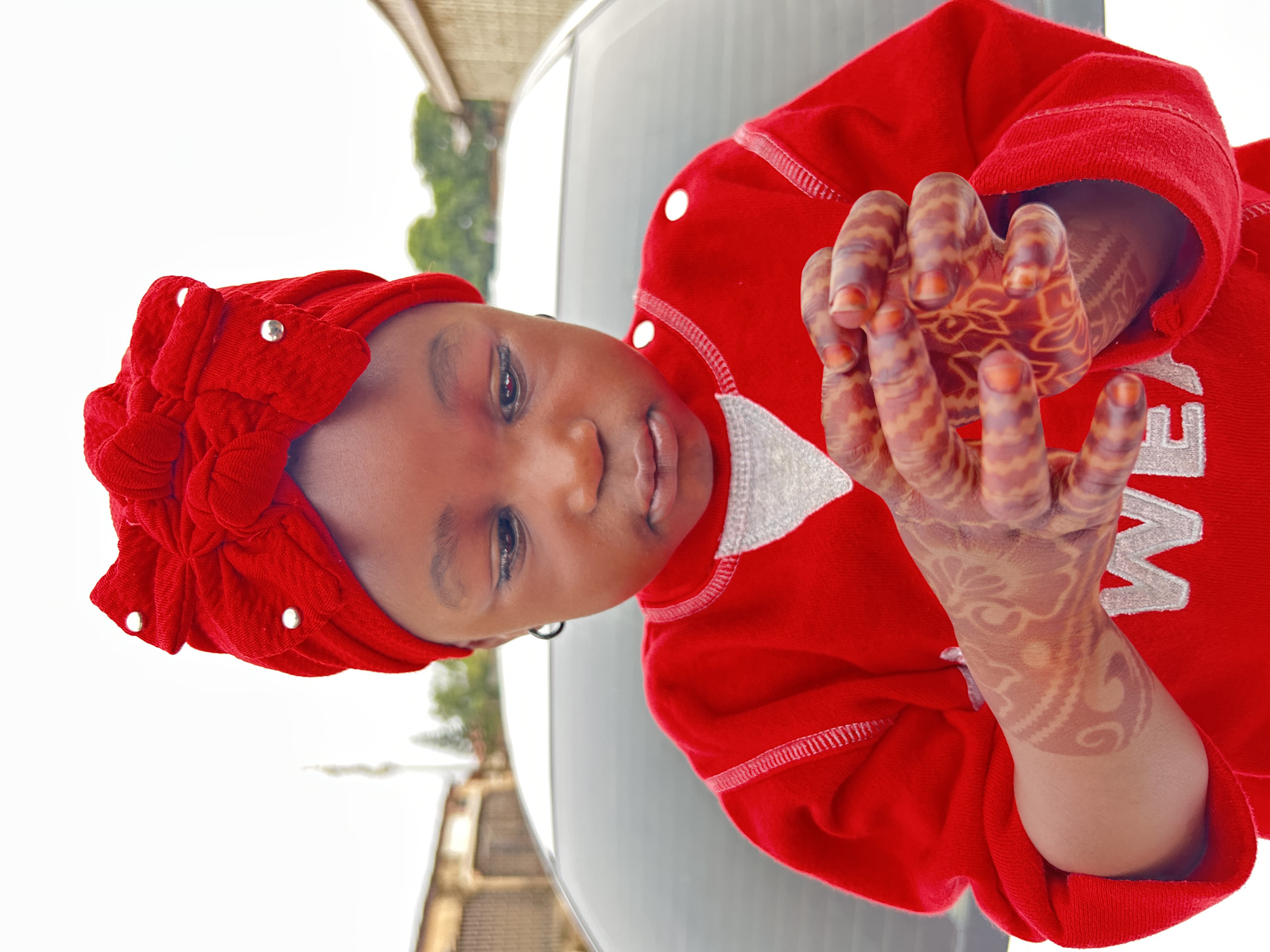 A young, dark-skinned child, possibly a girl, is wearing a red, button-down sweatshirt and a matching red head covering decorated with small, round, silver embellishments. The child is holding their hands together, palms facing each other, revealing intricate henna designs on the backs of the hands and fingers. The background is blurry, but suggests an outdoor setting. A silver car is visible behind the child and building. The scene is possibly set in Jos, Nigeria.