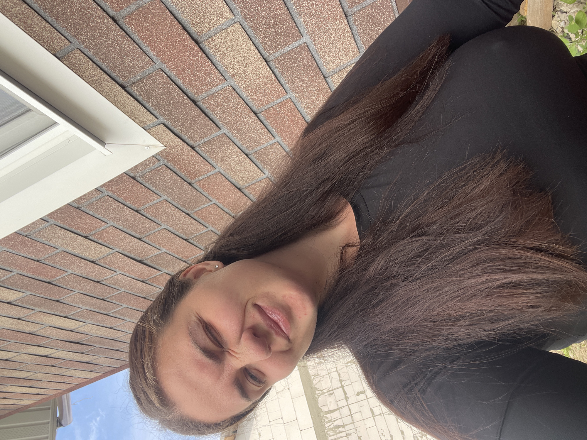 A young woman in a black long-sleeved top is posing in front of a brick wall and a white window frame. The brick wall has a red and brown pattern. The location is likely Мосметаллоконструкция Онт, Russia, based on the provided information. The woman is looking at the camera with one eye squinted. Her long, brown hair is draped over her shoulder. In the background, there is a glimpse of the sky and a building with white blocks.
