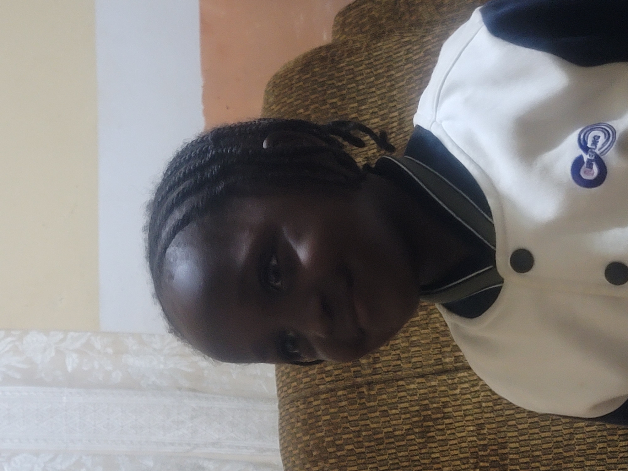A medium close-up shot captured in Barakin Akawo, Nigeria, depicts a young, dark-skinned child, likely female, with intricate dark braids styled on her head. The child is positioned slightly to the left of the frame, gazing upward and to the viewer's left with a subtle, closed-mouth smile and slightly puffed left cheek. She wears a two-toned varsity-style top: the main body and upper sleeves are white, while the lower sleeves and ribbed collar are dark blue or black. The collar features two thin horizontal stripes, one white and one light grey. Two large, circular dark buttons are visible on the front of the garment. An embroidered logo, featuring a blue stylized "C" with the word "STERLING" inside, intertwined with a blue infinity symbol, is present on the left chest area.
The child is seated against a light brown or ochre upholstered armchair or sofa, which exhibits a consistent, tightly woven geometric pattern of small rectangular or square shapes. The background on the left reveals a translucent white curtain with a subtle floral or patterned design, set against a pale yellow or cream wall. To the right, behind the armchair, a section of an orange-brown wall is visible, topped by a light beige or cream wall segment separated by a narrow white horizontal band. The lighting casts the child's face in shadow, particularly the left side, highlighting only certain features and the upper right portion of her forehead and shoulder, suggesting a light source positioned to the camera's right.