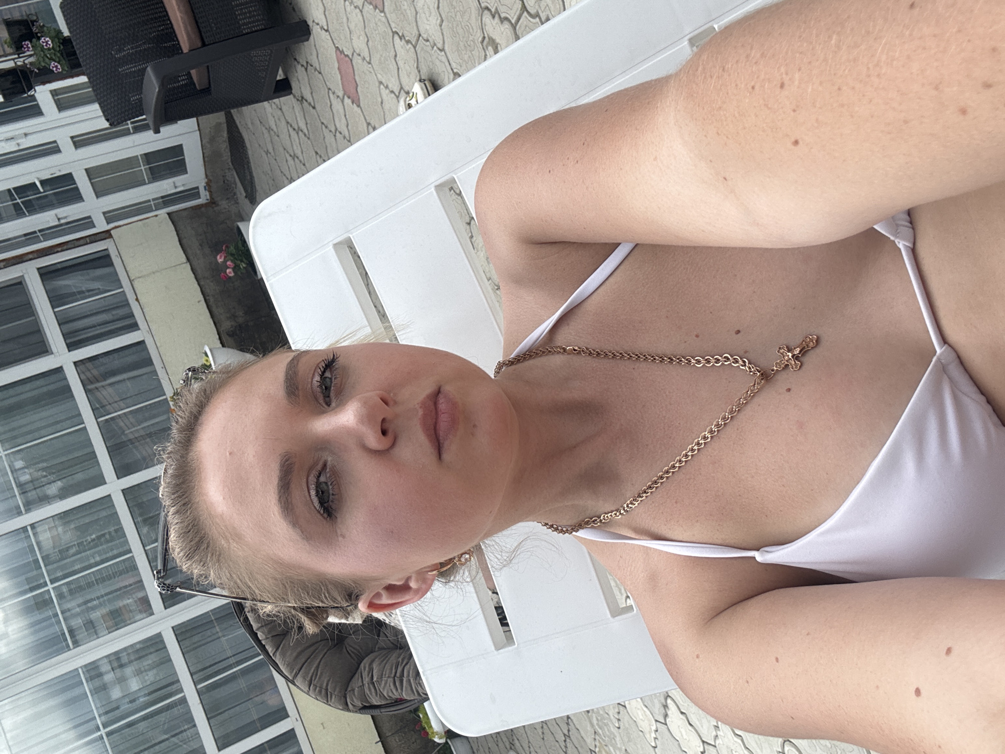 A young Caucasian woman with blonde hair pulled up in a bun and light makeup is sitting on a white lounge chair outdoors. She is wearing a white bikini top and a gold chain with a cross pendant. Sunglasses are perched on her head. The background shows a paved patio with a building and a dark wicker chair with a brown cushion. The image is a selfie, taken from a low angle.