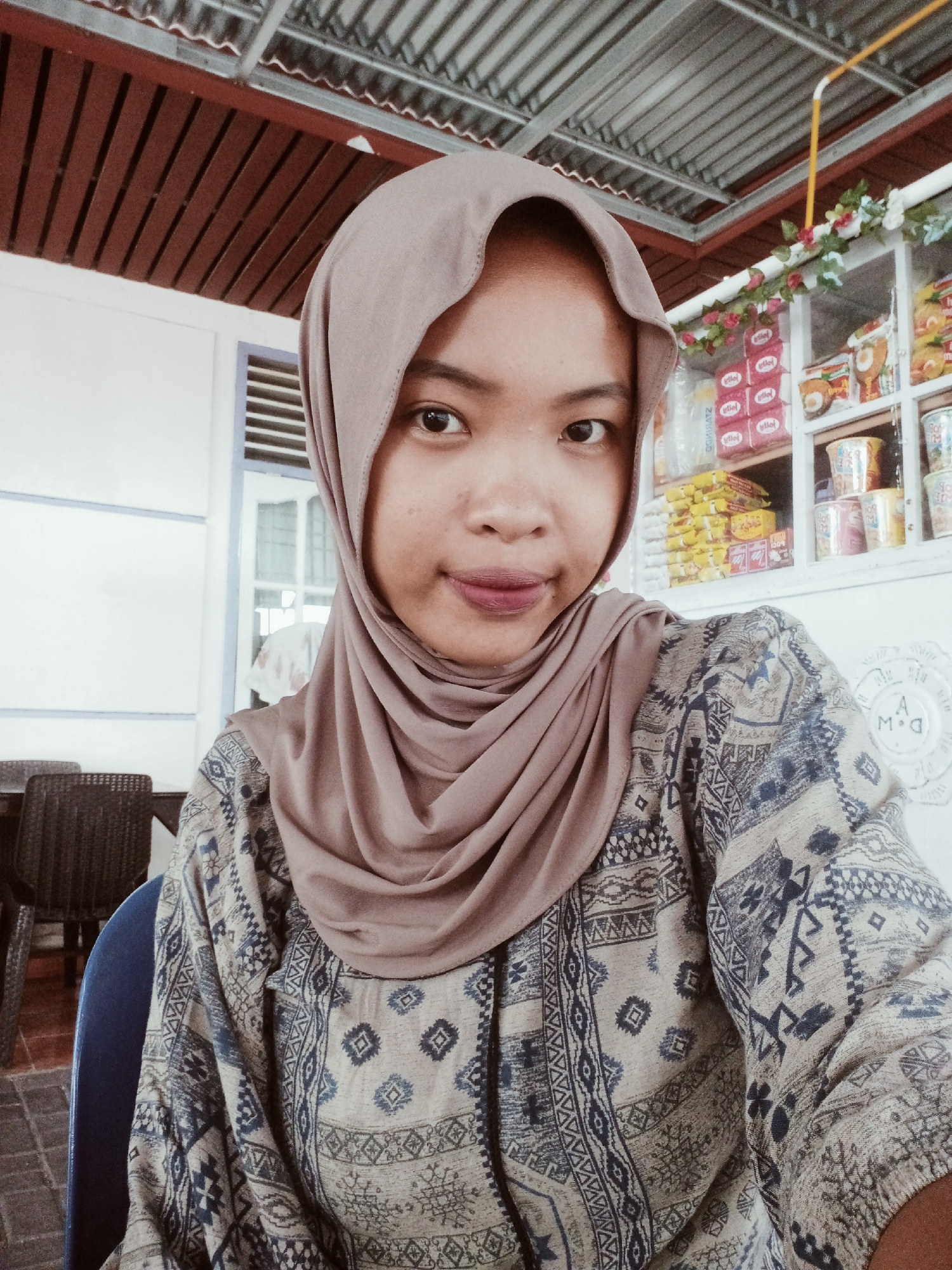 A medium shot shows a young woman wearing a tan hijab and a patterned long-sleeved shirt, seated in a cafe in Balik Alam, Indonesia. She is looking towards the camera with a neutral expression. In the background, shelves are stocked with various products, suggesting a small shop or convenience store attached to the cafe. A white wall and a corrugated metal ceiling are visible. Chairs and tables are partially visible in the foreground and left side of the image.