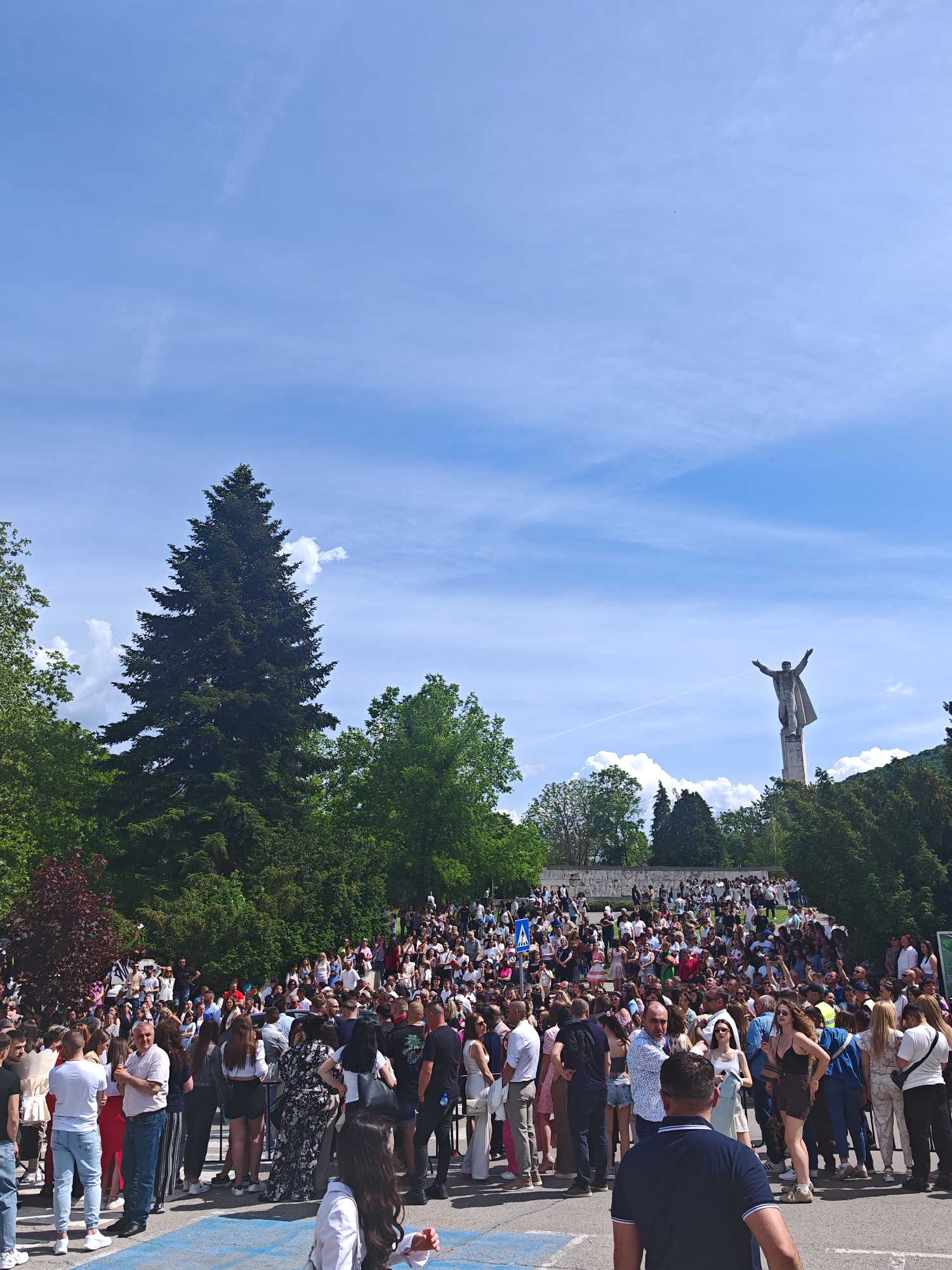 The image shows a large crowd of people gathered in Shumen, Bulgaria, in front of the Monument to 1300 Years of Bulgaria. The monument, featuring a figure with arms outstretched, stands in the background atop a hill. A tall evergreen tree is to the left. The sky is blue with scattered clouds. The crowd appears to be composed of people of varying ages and clothing styles, some wearing casual attire. The scene suggests a public gathering or event taking place at the monument site.