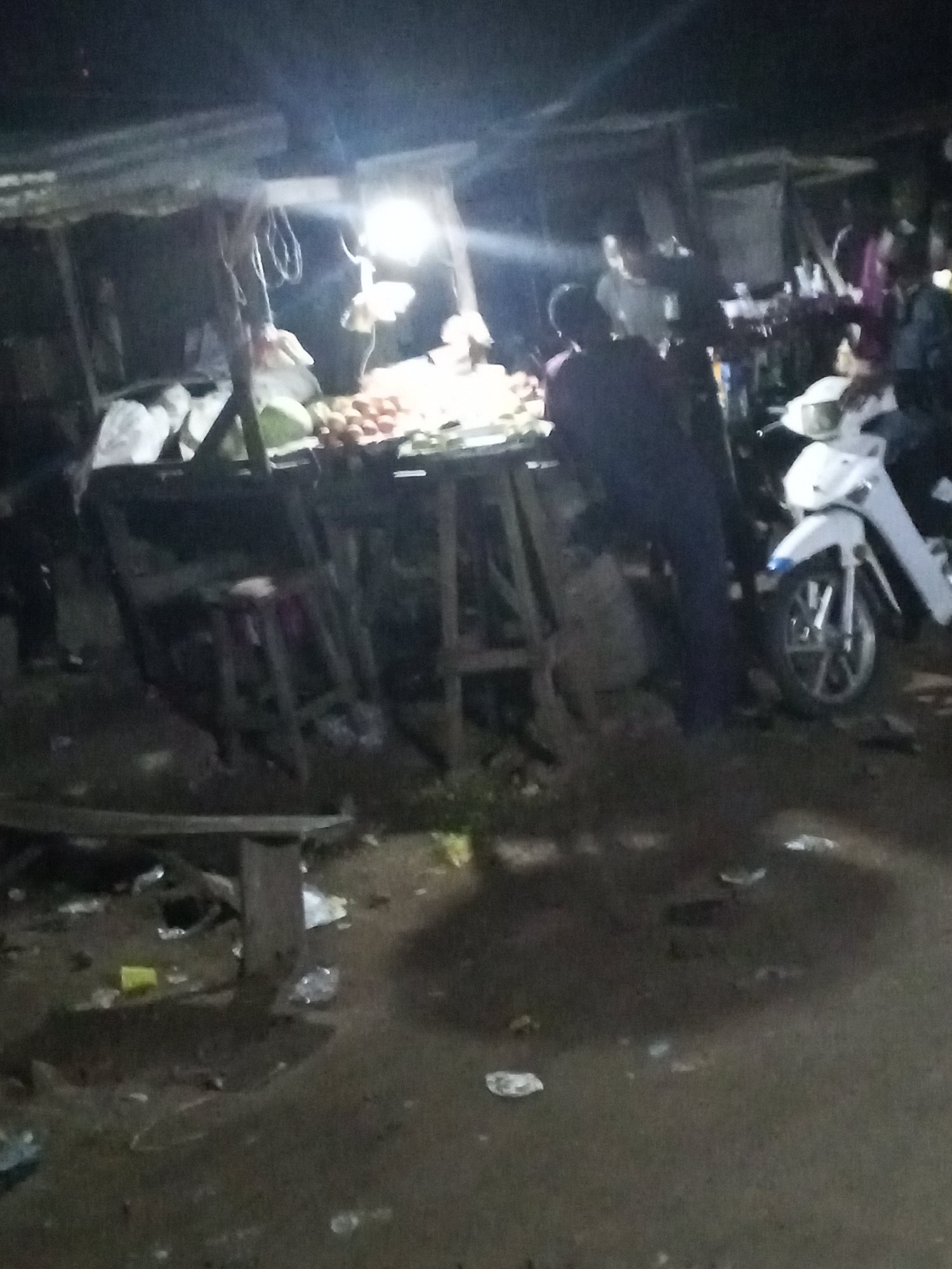 The image shows a nighttime scene in Lokoja, Nigeria, at an open-air market. A brightly lit stall displays produce, possibly fruits, under a makeshift awning. Several people are gathered around the stall, including at least one person in dark clothing standing near the stall and another person wearing a light-colored shirt in the background. A white motorcycle is parked near the stall. The ground appears to be littered with trash. The overall lighting is poor, making it difficult to discern details.