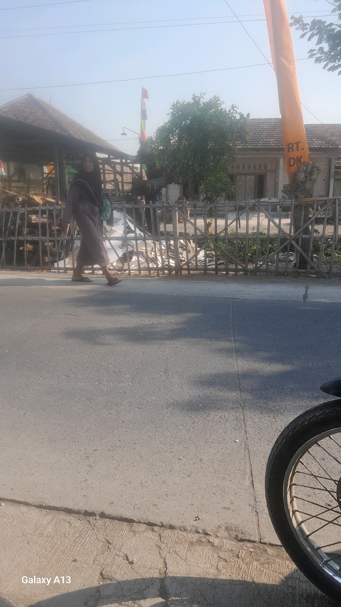 In Ngelo, Indonesia, a person wearing a dark hijab and a long, flowing garment walks along the side of a paved road. A green bag is slung over their shoulder. The person is walking away from the camera, towards the left side of the image. Behind the person, a rustic fence made of wooden poles is visible. Beyond the fence, there are buildings with tiled roofs, some appearing to be under construction. In the background, a tree with lush green foliage stands out. A tall, orange banner with dark lettering that reads "RT. DK." is attached to a tree stump to the right of the frame. The sky is clear and bright, indicating a sunny day. In the foreground, the wheel of a motorcycle is visible on the right side of the image. The road surface is paved and shows some cracks. The overall scene depicts a typical streetscape in a semi-rural or residential area.