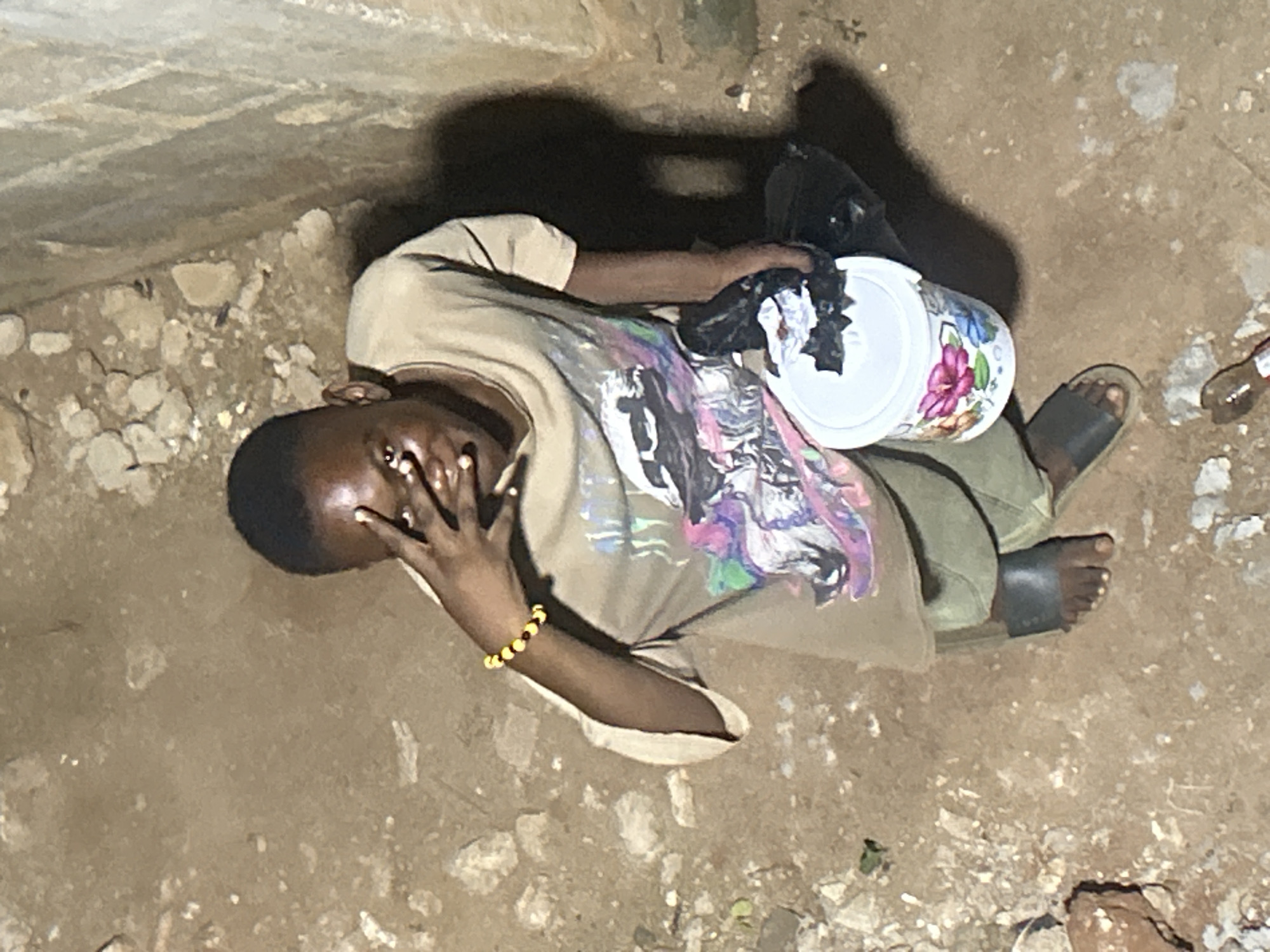 An overhead shot depicts a person lying on a dirt-covered ground. The person is wearing a light brown t-shirt with a colorful graphic, green pants, and black sandals. They are covering their face with their hand and have a yellow beaded bracelet on their wrist. A white bucket with a floral pattern sits beside them. To their upper right is a black plastic bag and a discarded bottle is to their lower right. The setting appears to be near a wall or structure with rough, uneven surfaces. The location is Unknown, Unknown.