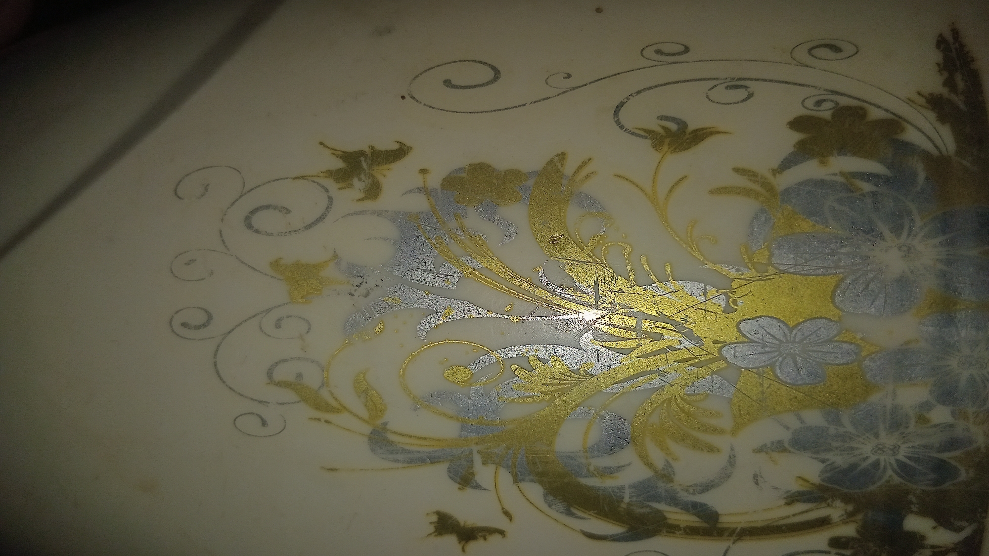 The image displays a close-up view of a decorative surface, likely a plate or a similar object, featuring an ornate floral pattern. The pattern is rendered in a metallic gold and silver finish against an off-white background. Swirling vines and leaves form the base of the design, interspersed with stylized flowers. Some of the flowers are depicted with delicate petals in silver, while others appear as golden outlines or filled shapes. The lighting in the image creates a slight glare in the center of the composition, highlighting the metallic elements of the decoration. The background is uniform and appears to be a smooth, solid surface. There are no people or other objects visible in the frame, and no specific location within a city can be determined from the image.