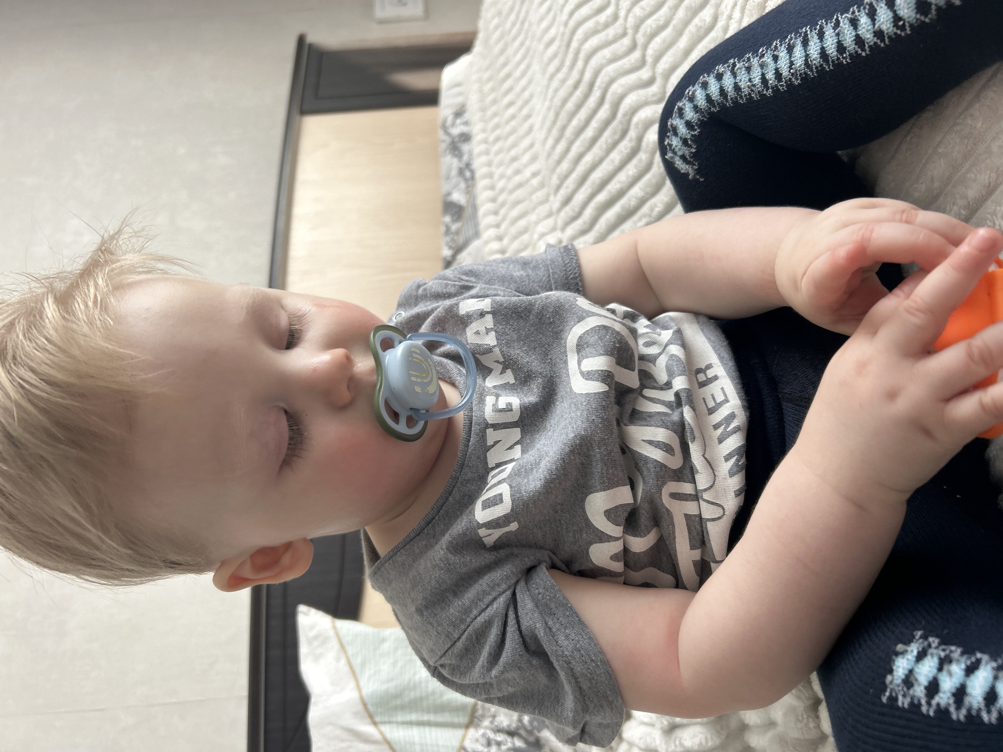 This image depicts a young child seated on a bed, potentially within a residential setting in the city of Chaikovsky, Russia. The child, appearing to be a toddler with light blonde hair and fair skin, has their eyes closed or directed downwards. A light blue pacifier with a white shield, featuring a blue 'U' design, is in the child's mouth. The child wears a grey short-sleeved t-shirt with white text, partially revealing "YOUNG MAN" and "RUNNER." Dark blue tights with a patterned light blue and white stripe along the outer seam cover the child's legs. The child's hands are clasped around a small, irregularly shaped orange object. The immediate environment includes a white, textured blanket or comforter on which the child sits. Behind the child, a dark wooden headboard is visible, and to the left, a white pillow with light blue and white stripes is partially present. The background wall is light-colored and unadorned.