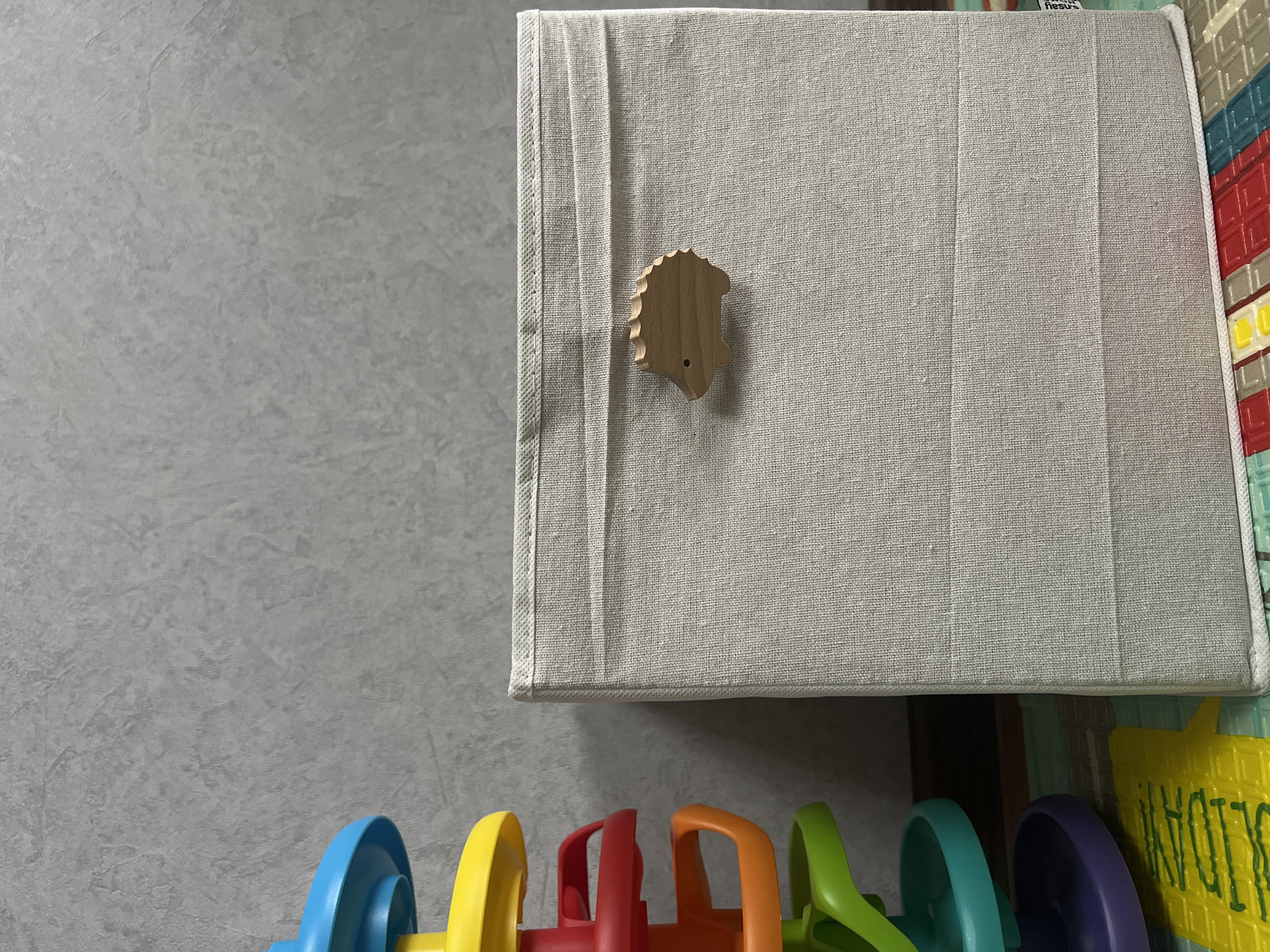 In an indoor setting, likely a child's room, a light gray textured wall serves as the backdrop. To the left, a brightly colored toy track with yellow, red, orange, green, blue, and purple sections is partially visible. To the right, a light beige fabric storage box stands upright. The box has a textured weave and features a small, light wood carving of a hedgehog attached to its front. The storage box rests on a colorful mat with geometric patterns in red, blue, and white. The scene is lit from the front, casting subtle shadows.
