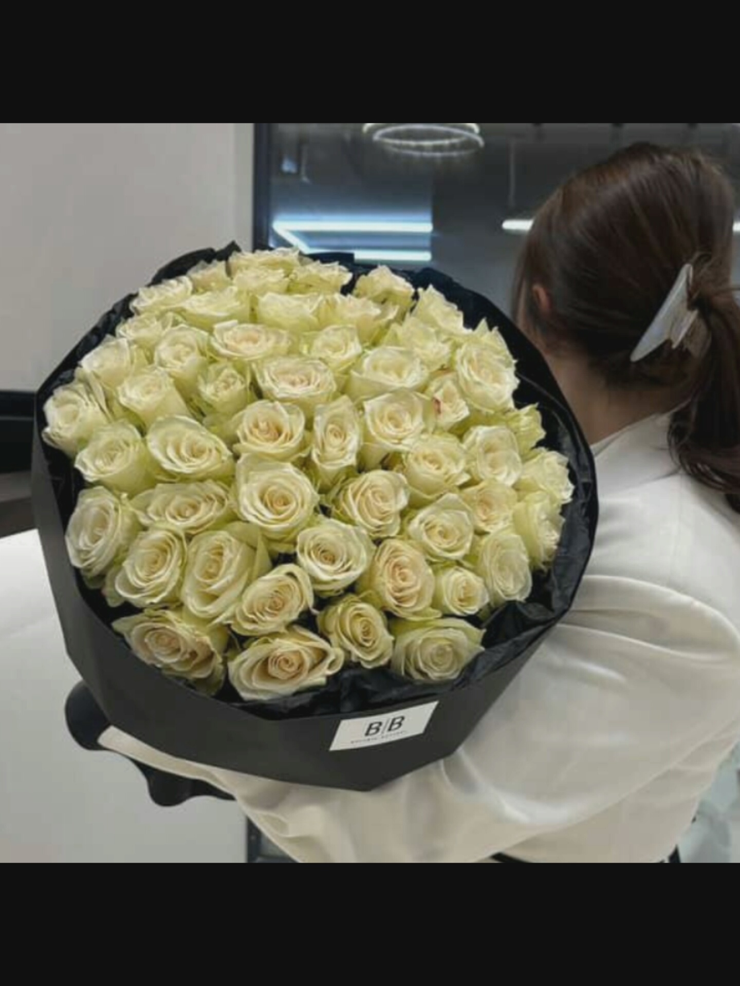 In Bucharest, Romania, a person is holding a large bouquet of white roses. The roses are tightly packed and arranged in a circular fashion, wrapped in black paper. A tag with "B B" is visible on the wrapping. The person's hair is dark and pulled back with a white clip. They are wearing a white garment. The background appears to be an indoor setting with modern lighting.