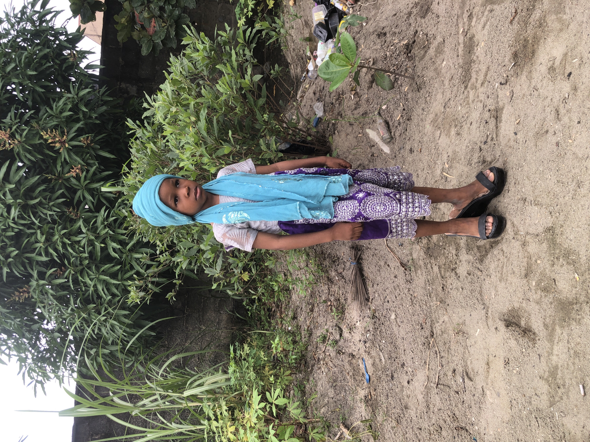 A young girl stands facing forward in a dusty, outdoor environment. She is wearing a light grey shirt with a silver patterned design, purple pants with a white patterned trim, and a turquoise headscarf and matching shawl. She is also wearing black wedge sandals. Behind her, there is a concrete wall and lush green vegetation, including tall grass and trees. The ground is covered in dirt and sand, with scattered debris including a broom, a blue plastic spoon, and various pieces of trash. The scene appears to be in Ado, Nigeria.