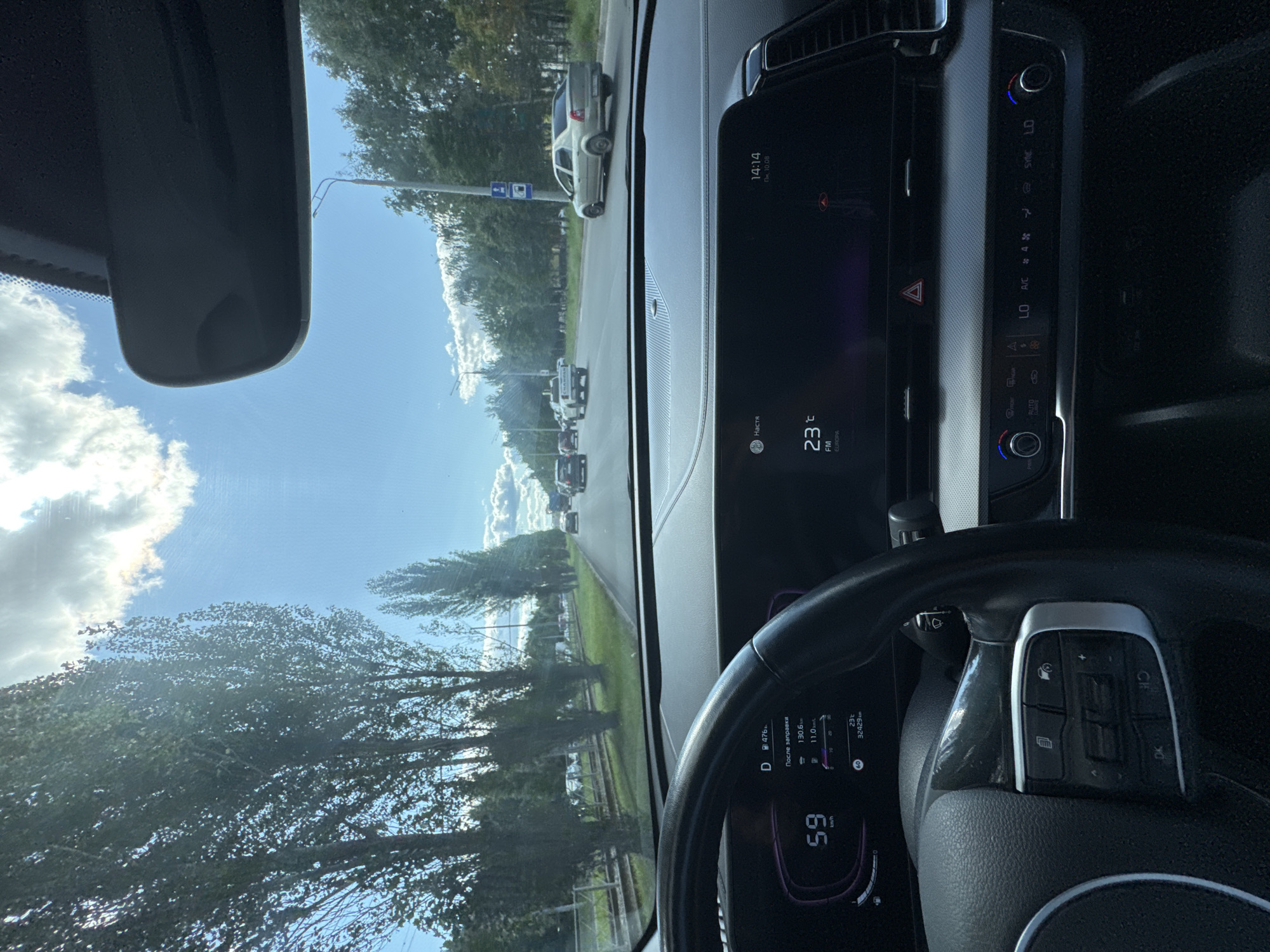 The image shows the interior of a car, looking out through the windshield at a street scene in Kazan, Russia. The time displayed on the car's dashboard is 14:14. The exterior temperature is 23 degrees Celsius, and the date is October 8th. The car is traveling at a speed of 59 km/h. The dashboard indicates that 130.6 km have been driven since the last refueling, with an average fuel consumption of 11.0 km/l. The odometer shows 32,429 km. The road is lined with trees, and there are other cars on the road, including a silver sedan and a white pickup truck. The sky is blue with some clouds. The interior of the car includes a steering wheel and a digital display.