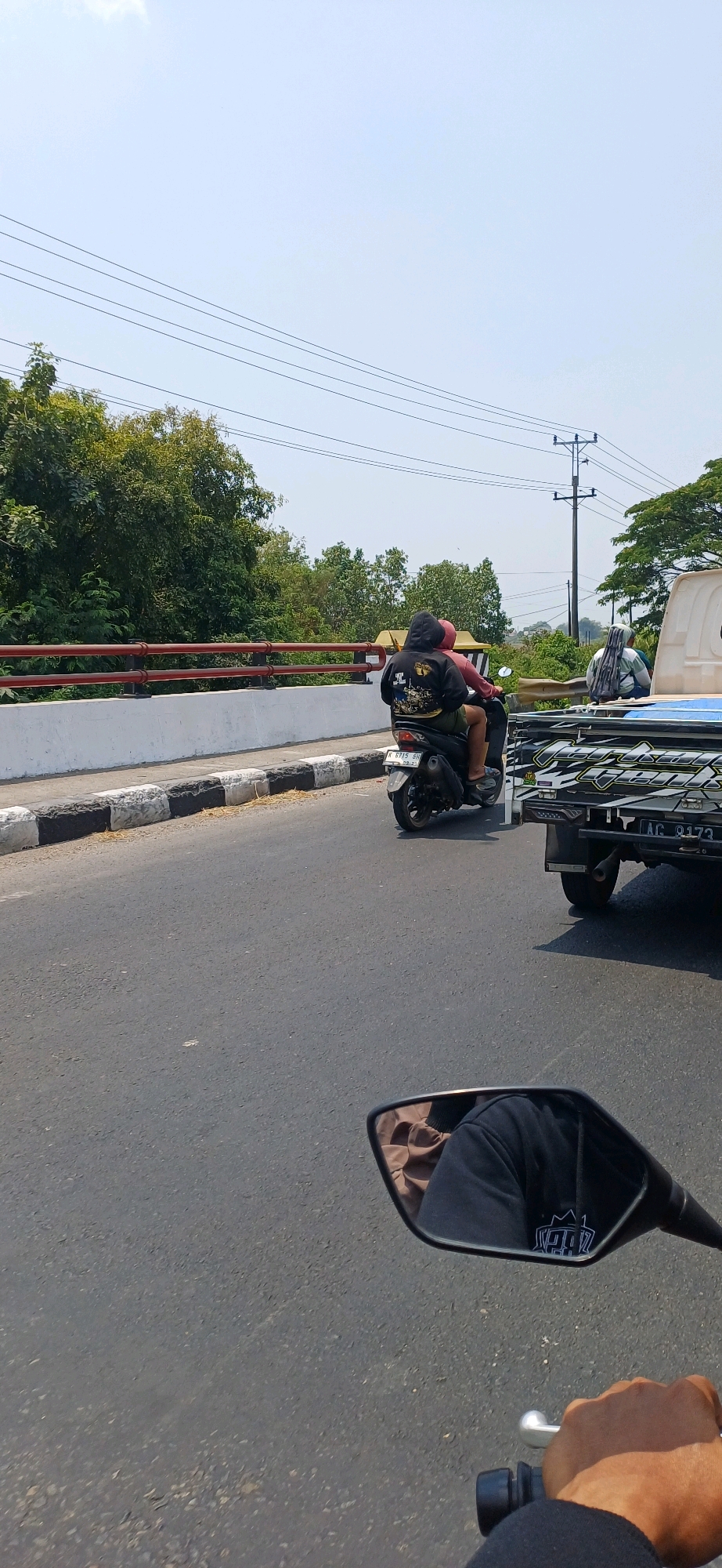 In a street scene in Kecamatan Pati, Indonesia, a motorcycle carries two individuals. The person in the front, wearing a dark hoodie with a graphic design, is operating the motorcycle. The person in the back, wearing a red shirt, is seated behind them. They are traveling on an asphalt road with a white and black striped curb on the left. A concrete barrier with a red railing runs along the side of the road, separating it from a tree-lined area. In the background, a utility pole with multiple wires is visible. To the right of the motorcycle, a white truck with a blue tarp-covered cargo bed is also on the road. The scene is under a clear blue sky with a bright sun. A motorcycle's rearview mirror in the foreground reflects a person's arm and shoulder wearing dark clothing and a tan garment.