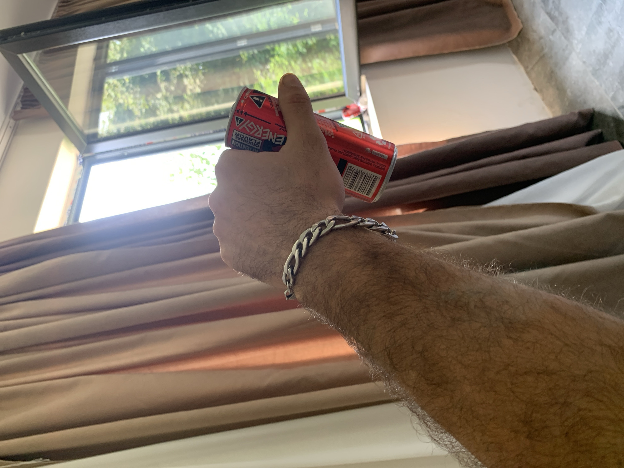 An arm with a silver bracelet is holding a red energy drink can in front of a window with light brown curtains. The can has a label with the word "ENERGY" in white letters. The background shows trees and greenery outside the window. The scene appears to be indoors, possibly a room with a view of nature. The specific location in the city of Kakhati, Georgia, is not identifiable from the image.